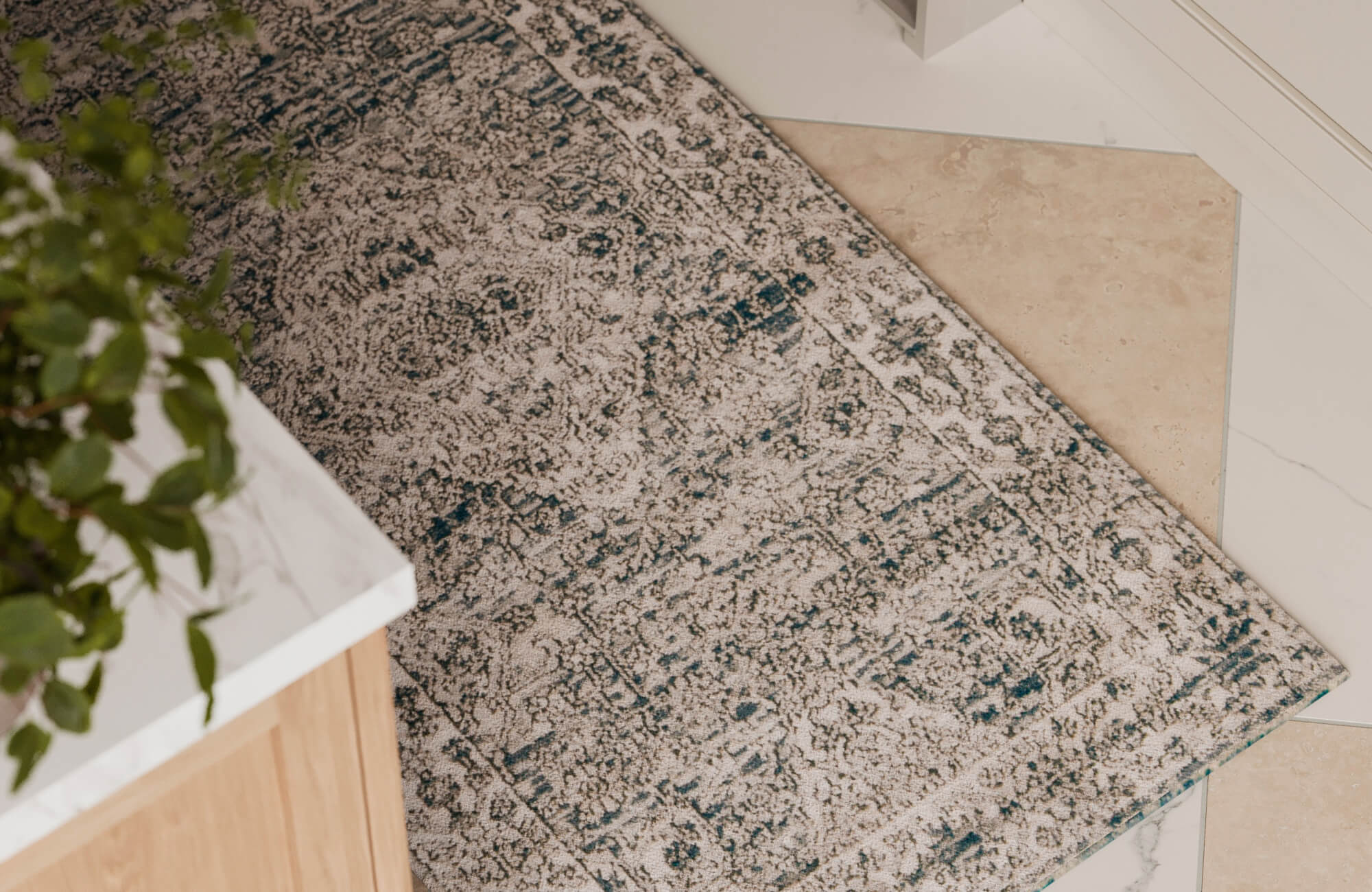 Corner view of a distressed-style rug in muted beige and teal tones, adding texture atop a tiled kitchen floor.