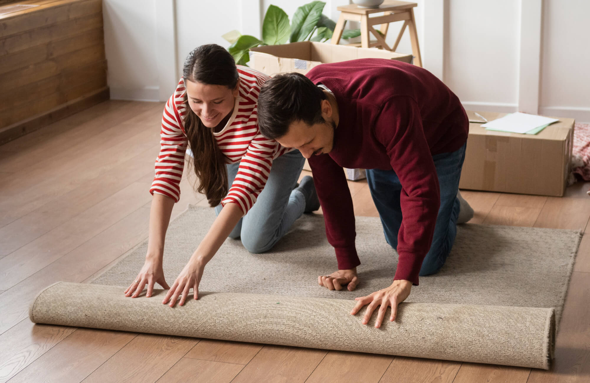 null Smiling couple rolling out a neutral-toned polypropylene rug over hardwood floors in a cozy, light-filled room with moving boxes.