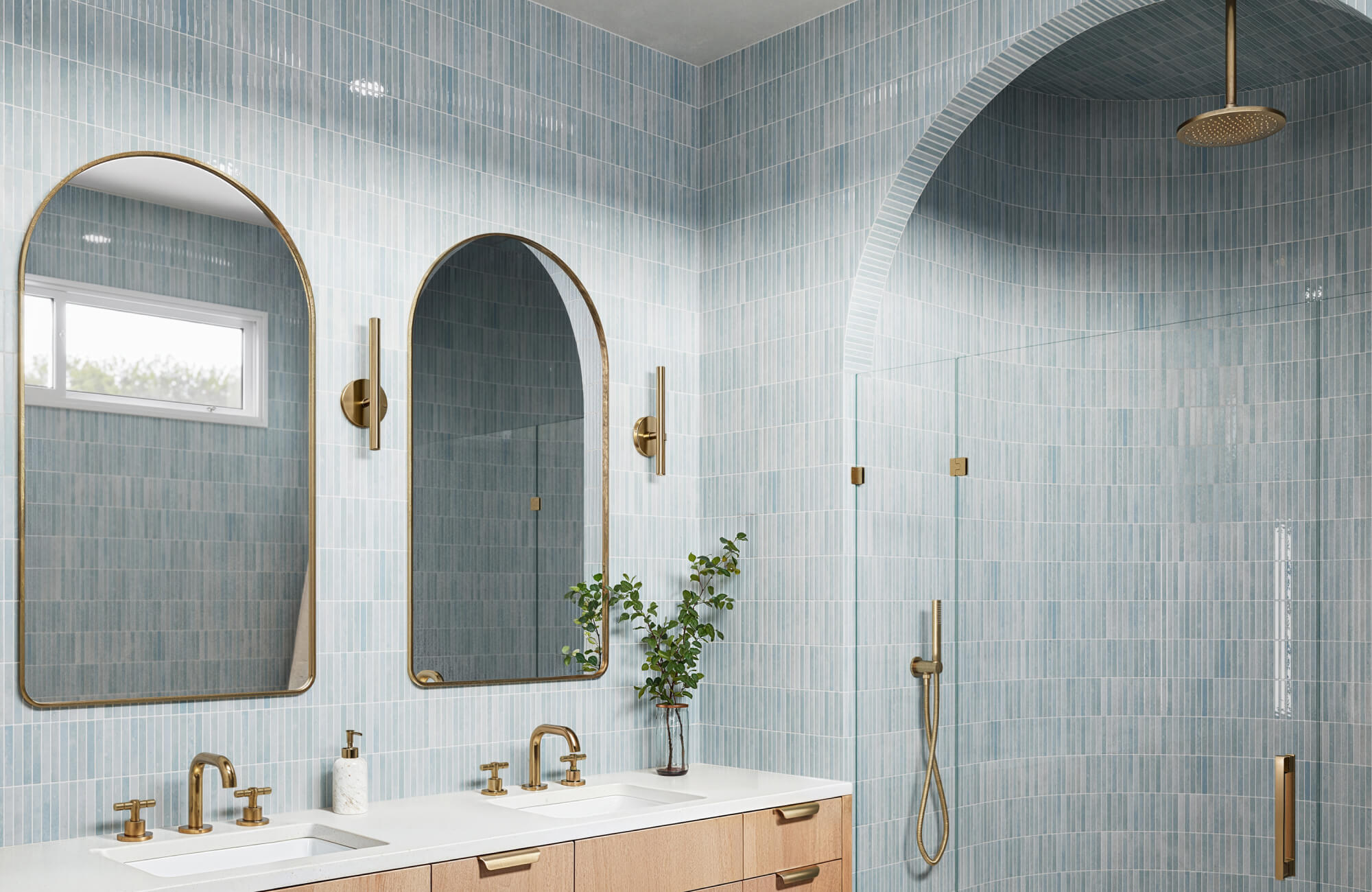 null A serene bathroom with soft blue vertical subway tiles, brass fixtures, arched mirrors, a vanity, and a walk-in shower with matching arched detailing.
