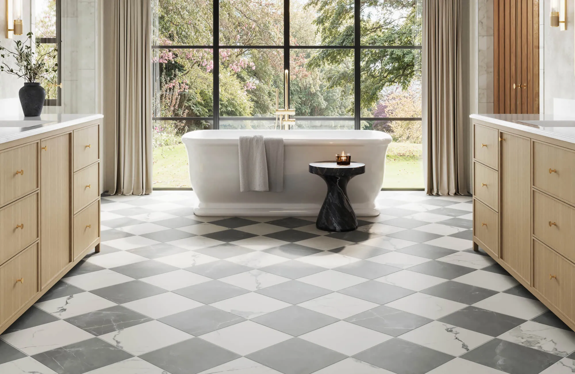Bright bathroom with a freestanding white bathtub set before floor-to-ceiling windows, featuring 12x12 checkerboard floor tile in soft gray and white, light wood vanities, brass fixtures, beige curtains, and garden views.