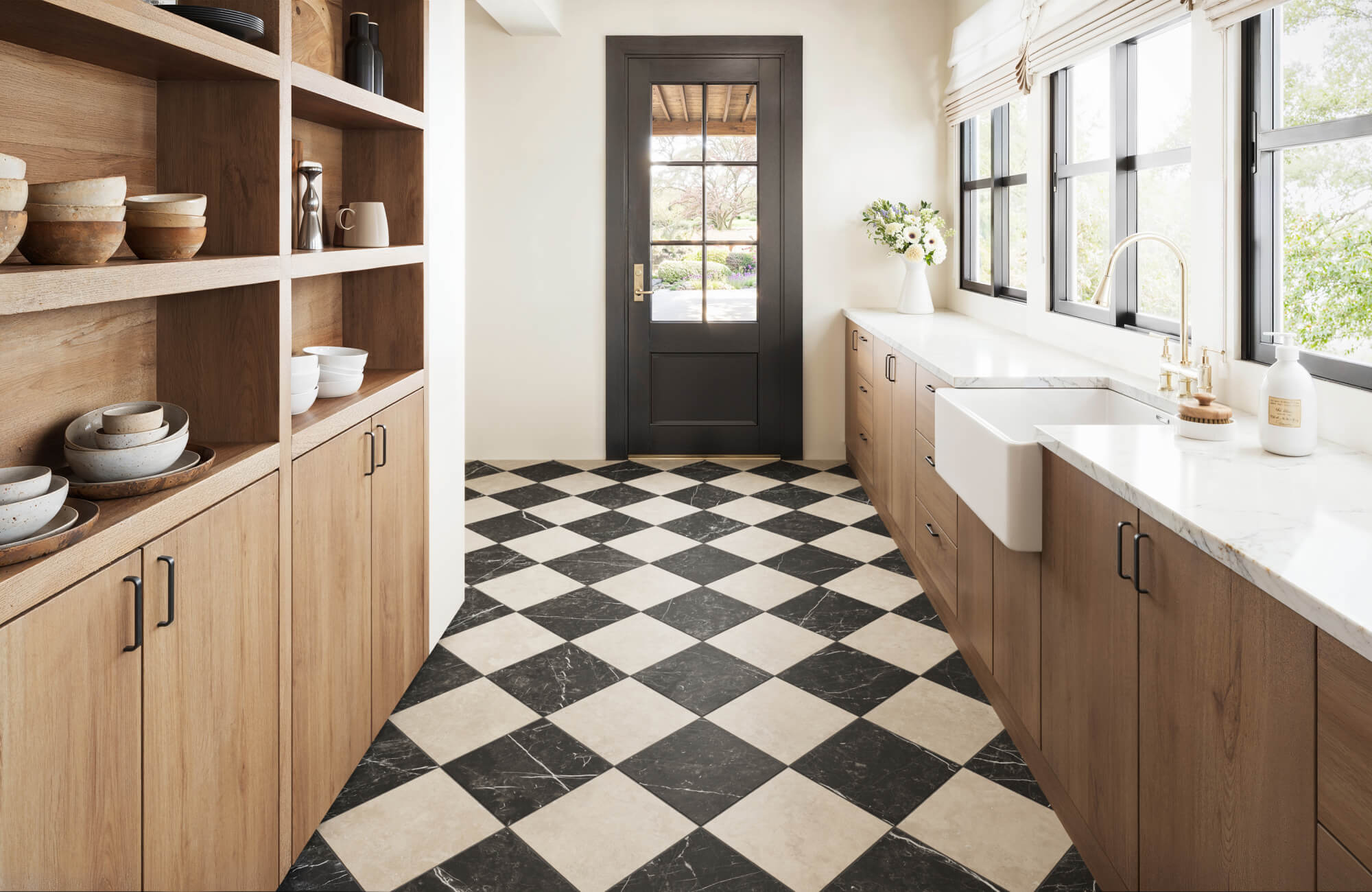 null Sunlit kitchen with 12x12 black and warm beige checkerboard floor tile, light wood cabinetry, open dish shelving, white countertops, a farmhouse sink, black-framed windows, and a glass-paneled back door.