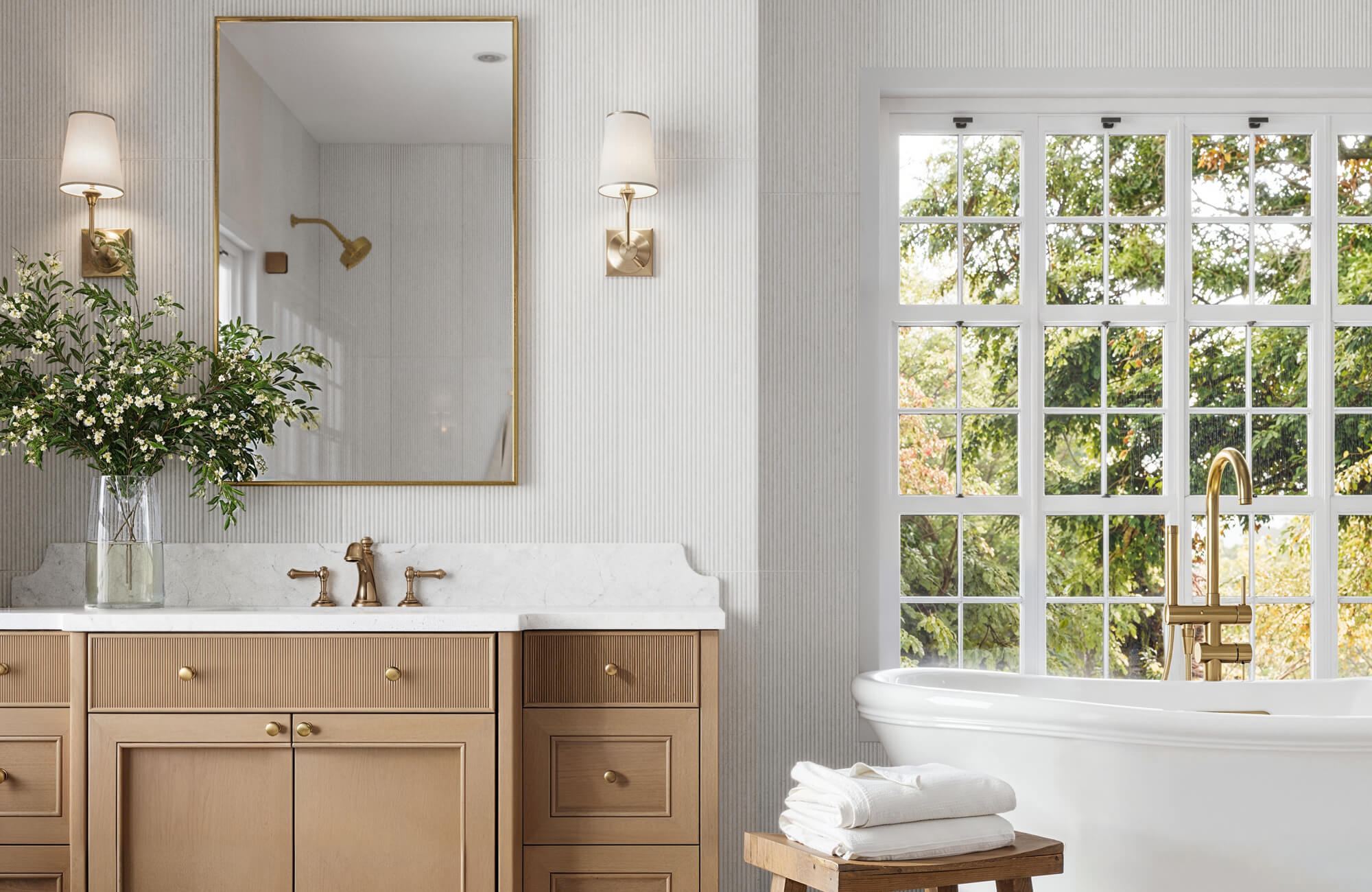 Bright bathroom with beige cabinetry, marble countertop, brass fixtures, and a large window overlooking lush greenery. Freestanding tub adds elegance.