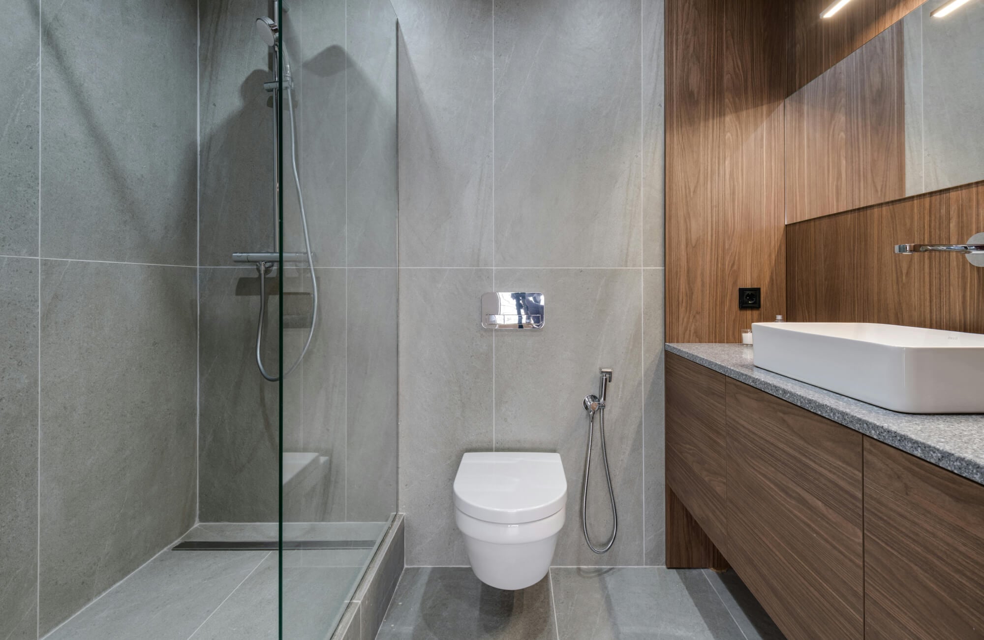 Sleek modern bathroom with large gray wall and floor tiles, a floating toilet, a glass-enclosed shower, and a warm wood-paneled vanity for contrast.