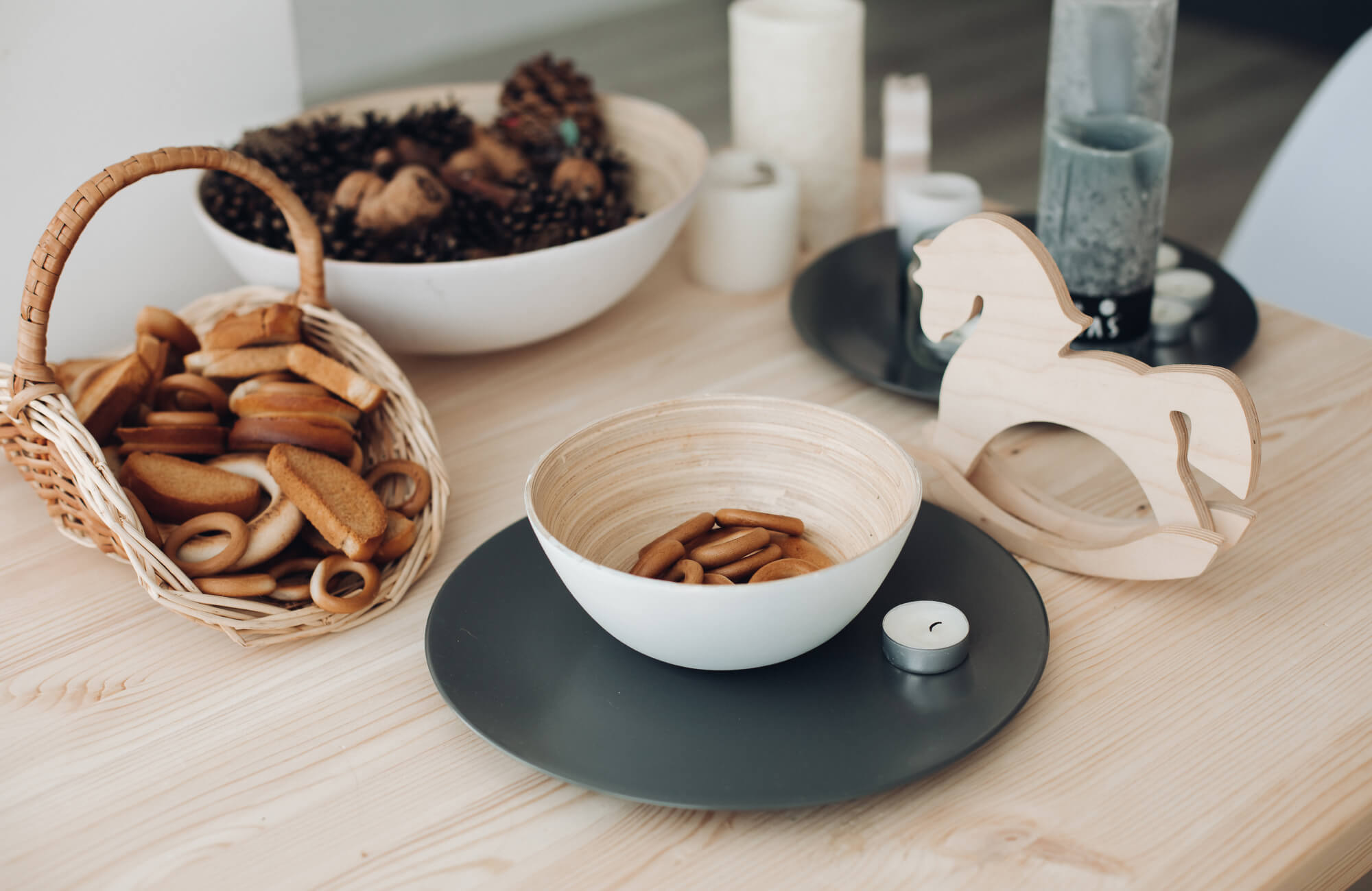 A decorative bowl filled with wooden rings sits on a tabletop alongside woven baskets, pinecones, candles, and wooden accents, creating a warm, nature-inspired display.