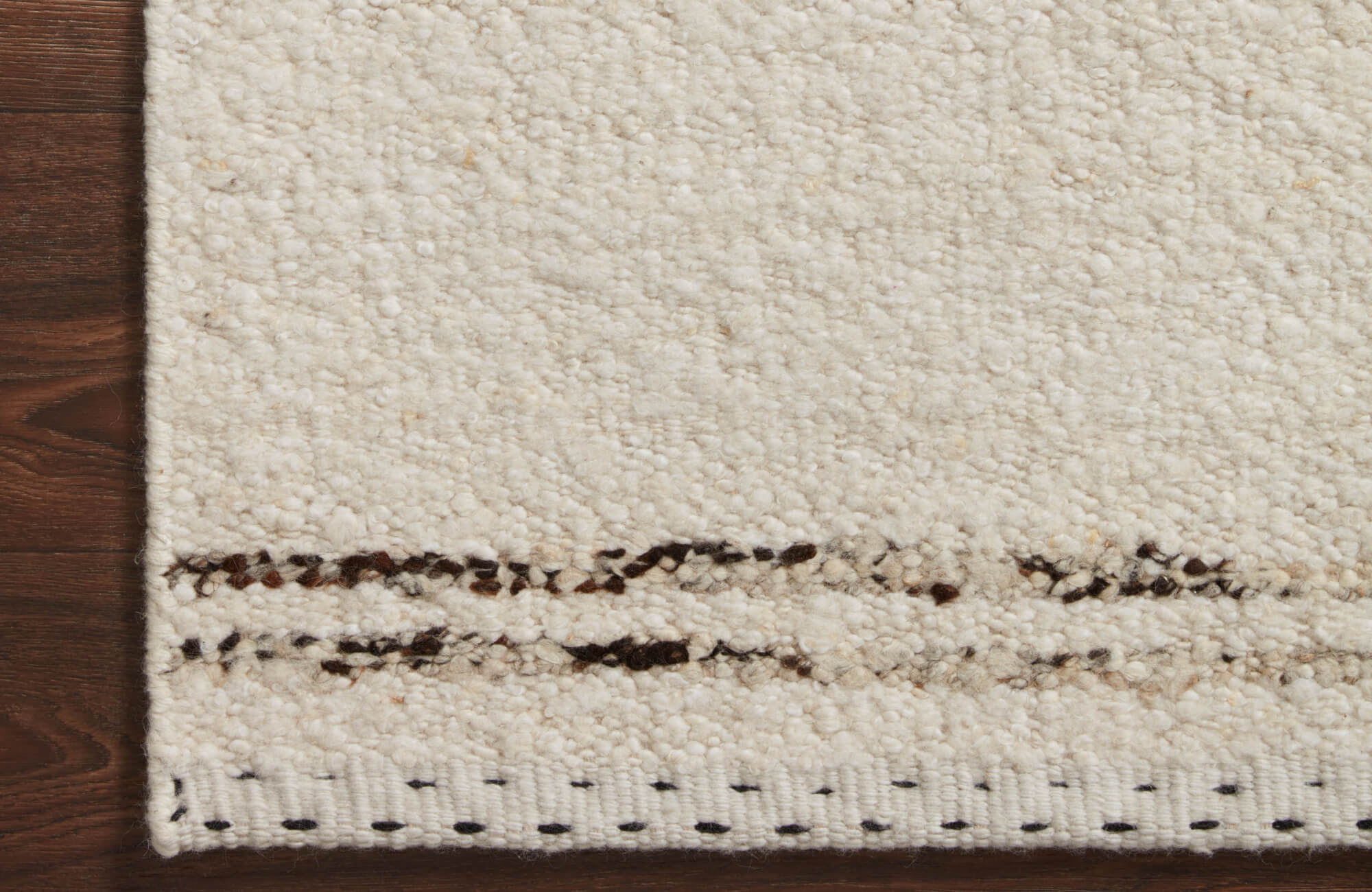 Close-up of ivory wool rug with dense nubby texture, subtle tonal variation, and stitched edge detail on dark wood flooring.