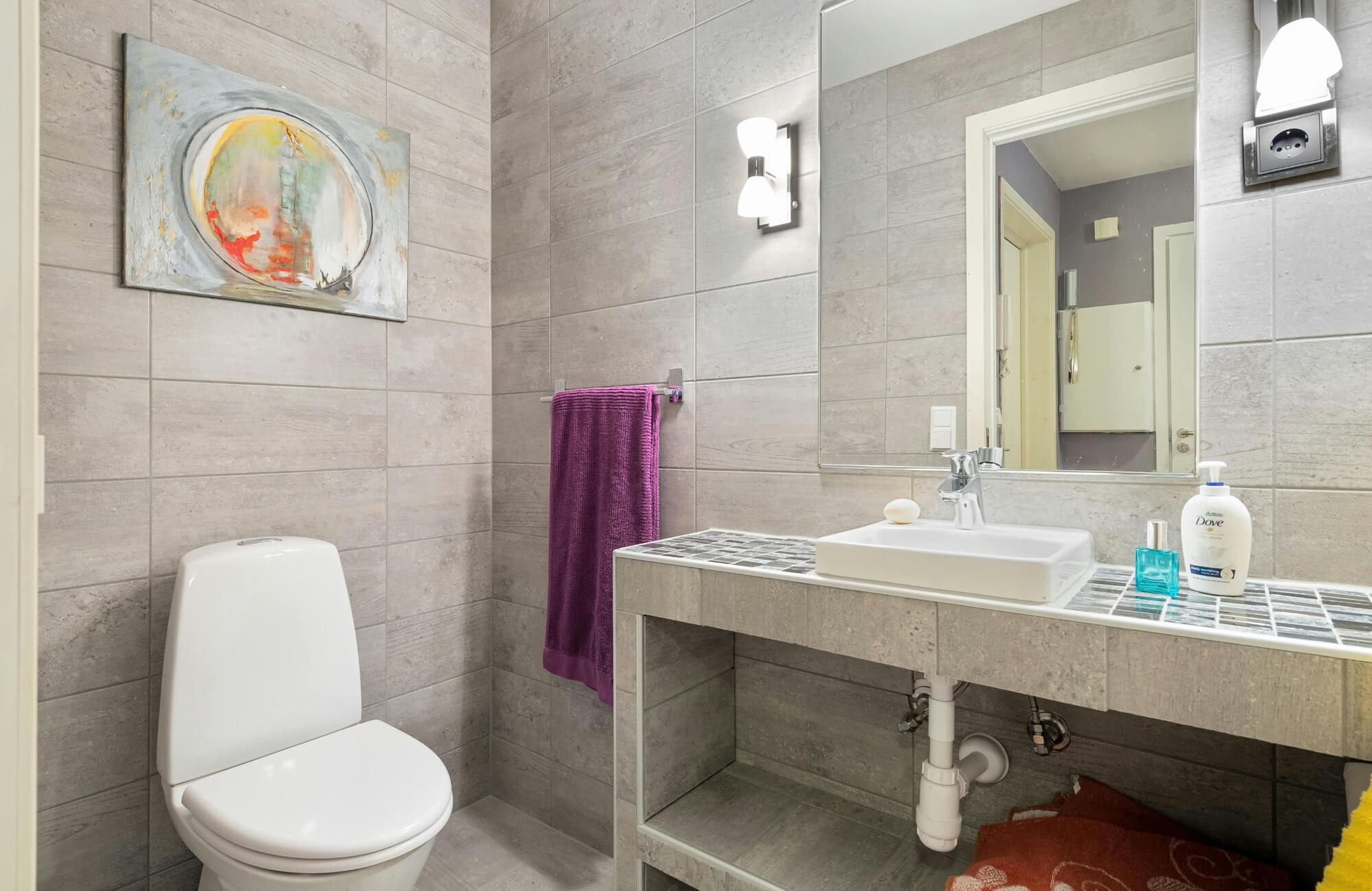 null Compact bathroom with gray tiled walls, wall-mounted sink, mirror, purple towel, and small abstract painting above toilet.