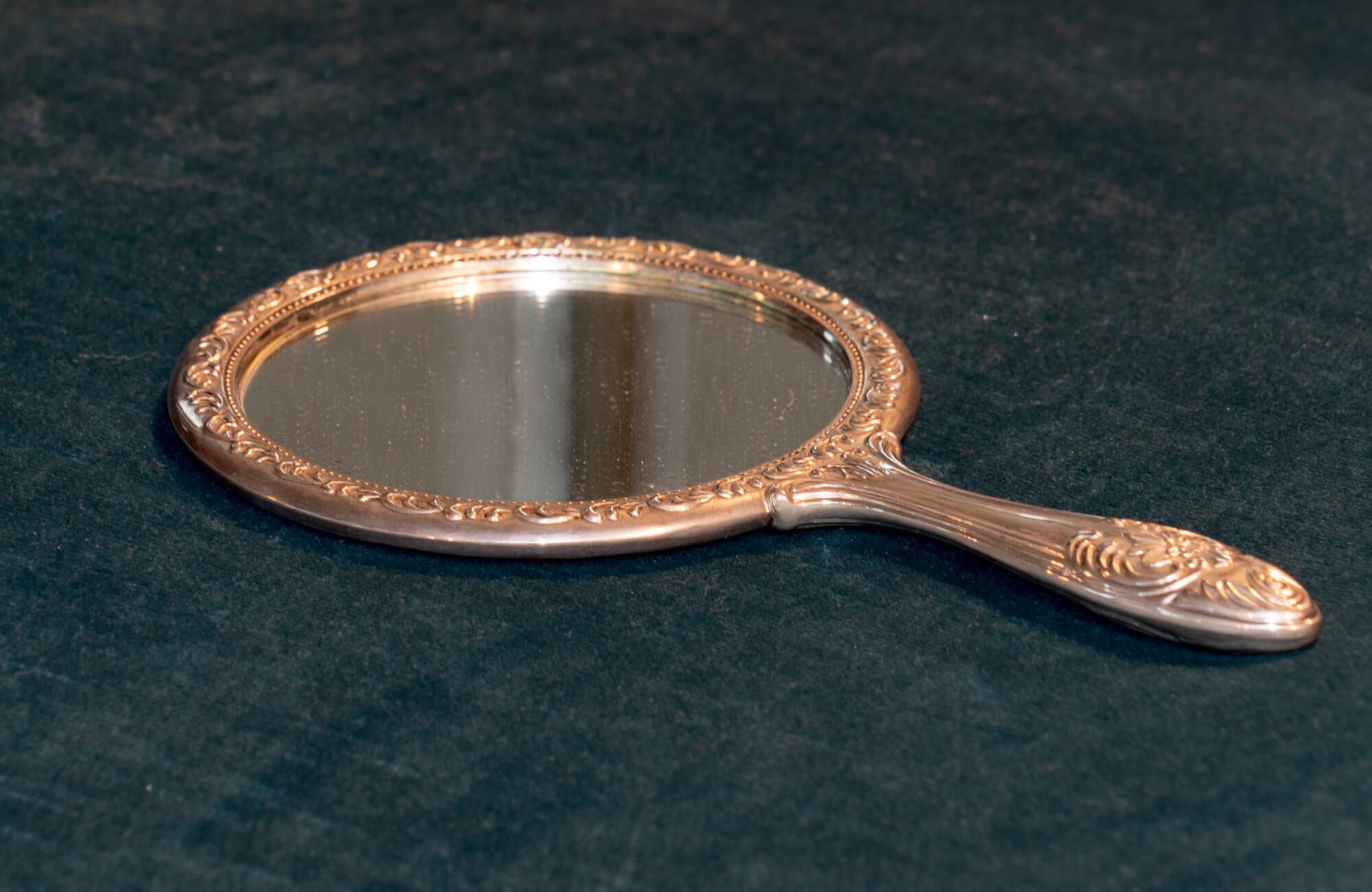 Ornate vintage handheld mirror with an embossed metal frame and handle, resting on a dark velvet surface.