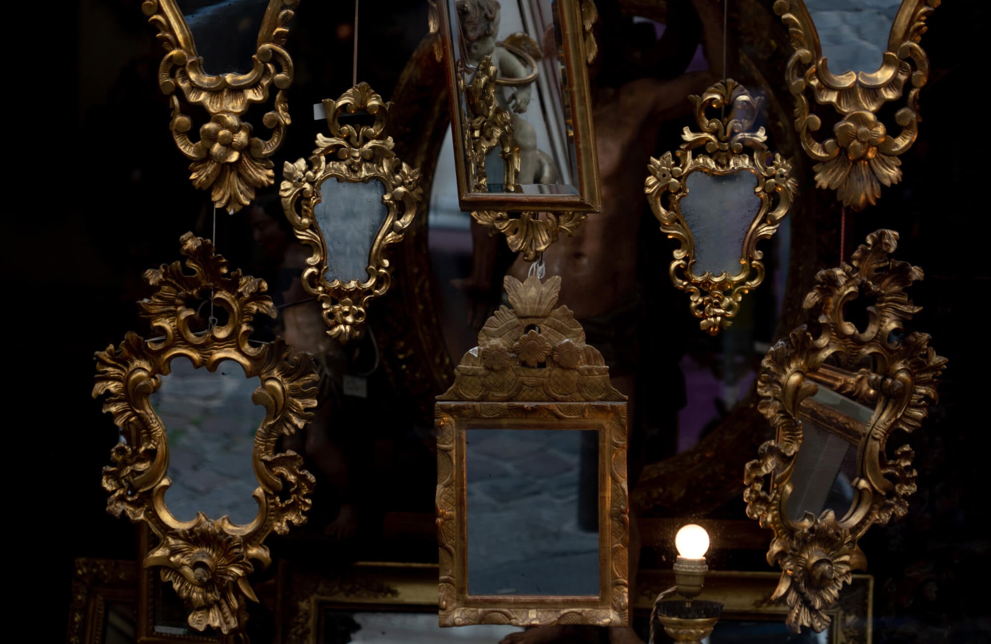 Collection of ornate antique mirrors with gilded frames displayed against a dark background, reflecting sculptures and ambient light.
