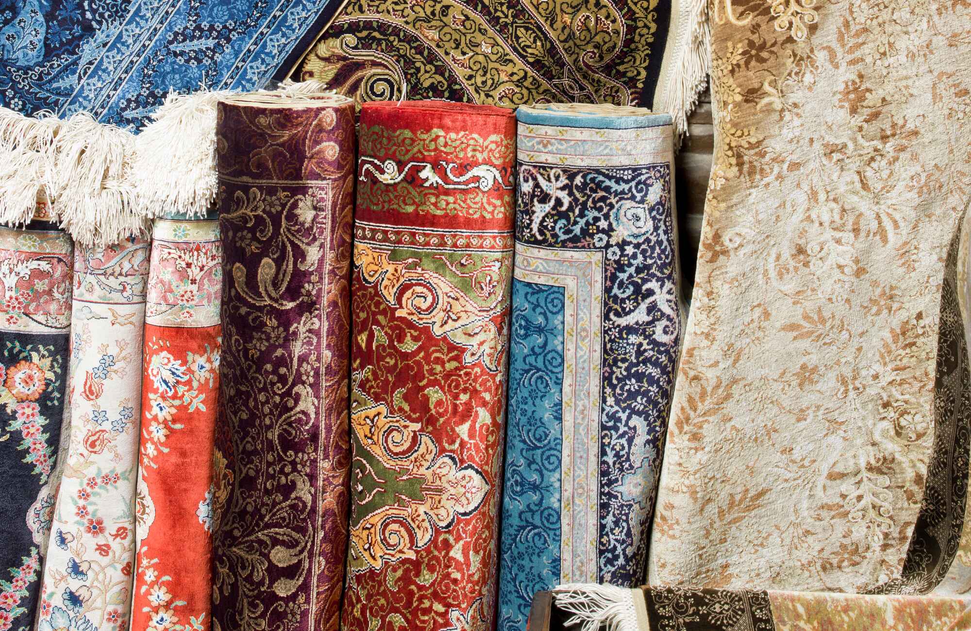 null Multiple upright rolled rugs in bold hues like crimson, navy, and ivory, each featuring detailed floral and border patterns with visible fringe ends.