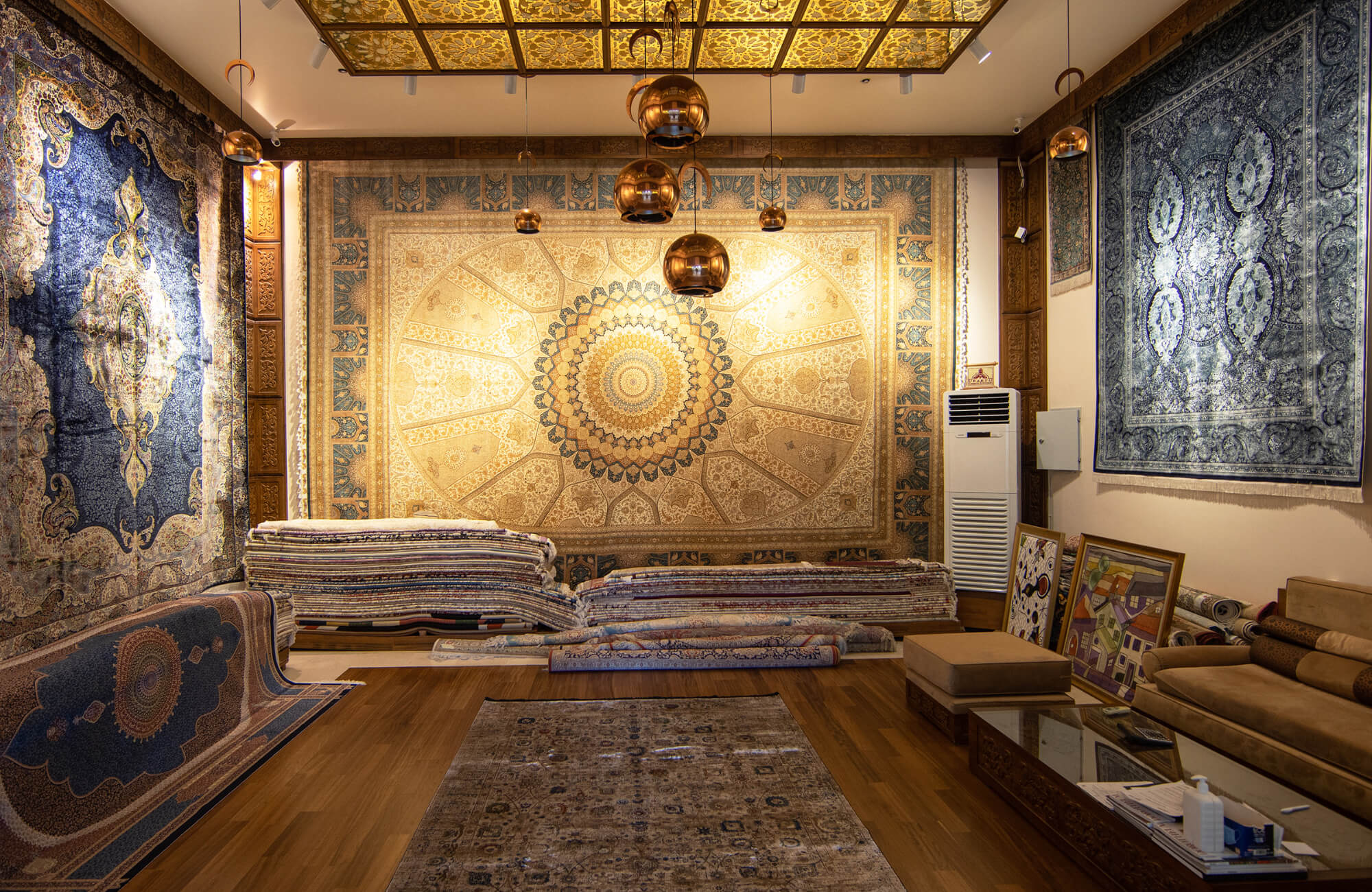 null An upscale rug showroom with large wall-mounted carpets showcasing intricate medallion designs, stacks of folded rugs, and warm pendant lighting overhead.
