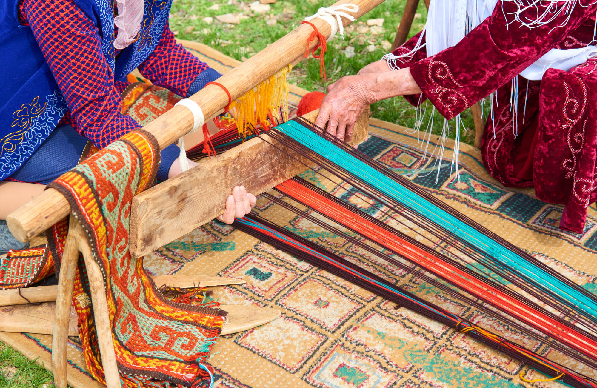 null Two artisans weaving a patterned rug on a wooden loom, guiding brightly colored threads in red, teal, and yellow into a geometric textile.