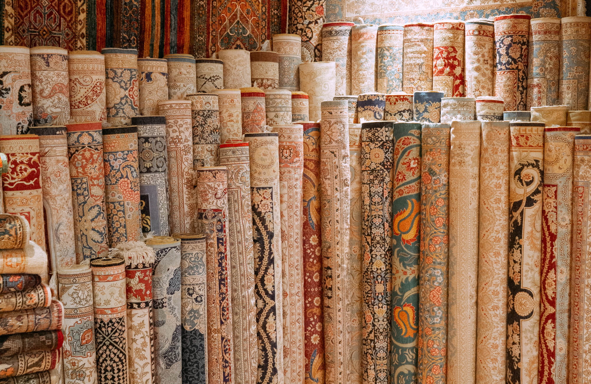 null A tightly packed display of rolled rugs in soft beige, blue, and rust tones, featuring ornate floral, vine, and medallion designs in a retail setting.