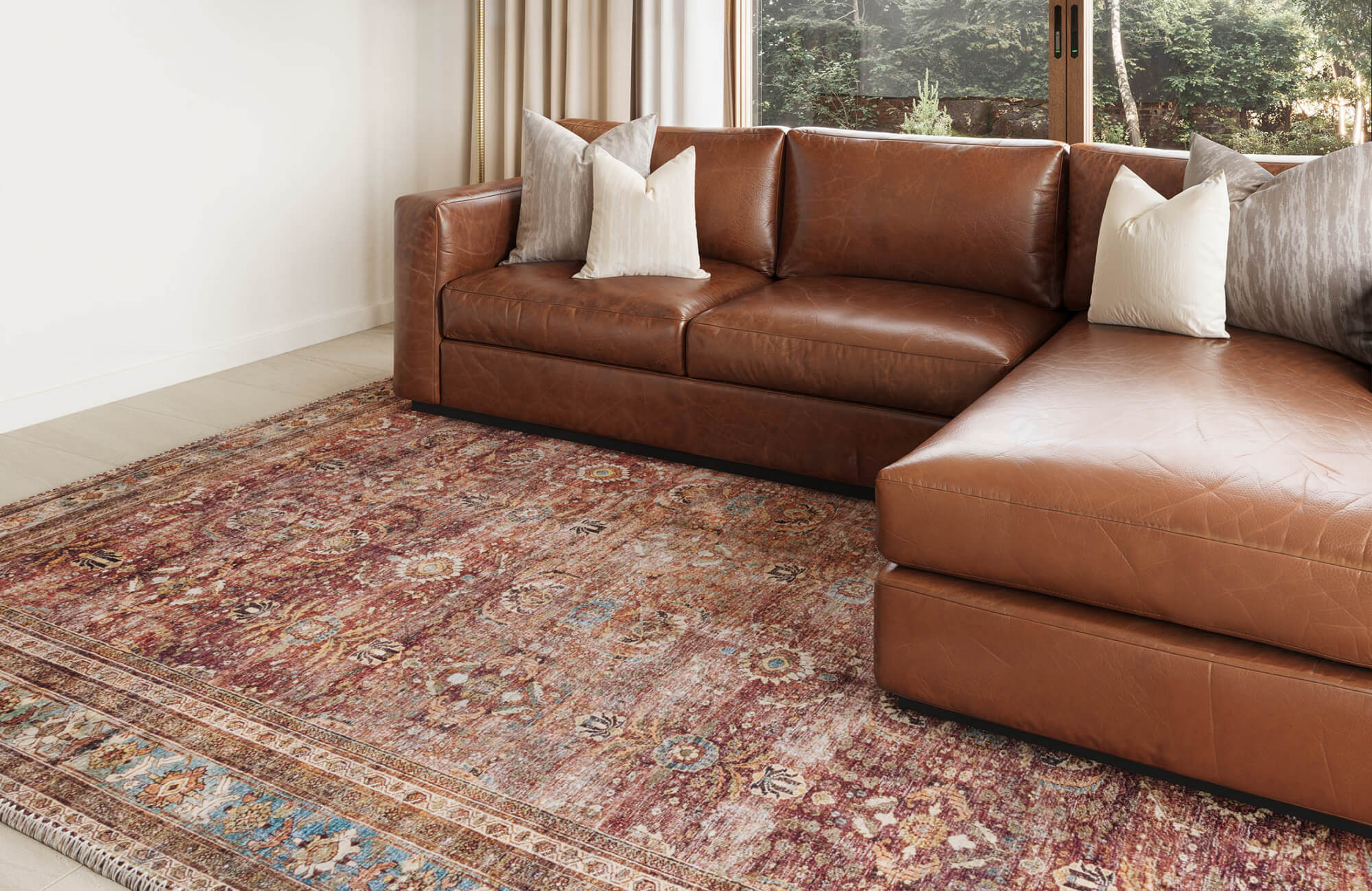 null A distressed vintage-style rug in burgundy and faded blue tones beneath a brown leather sectional, styled in a sunlit living room with neutral finishes.