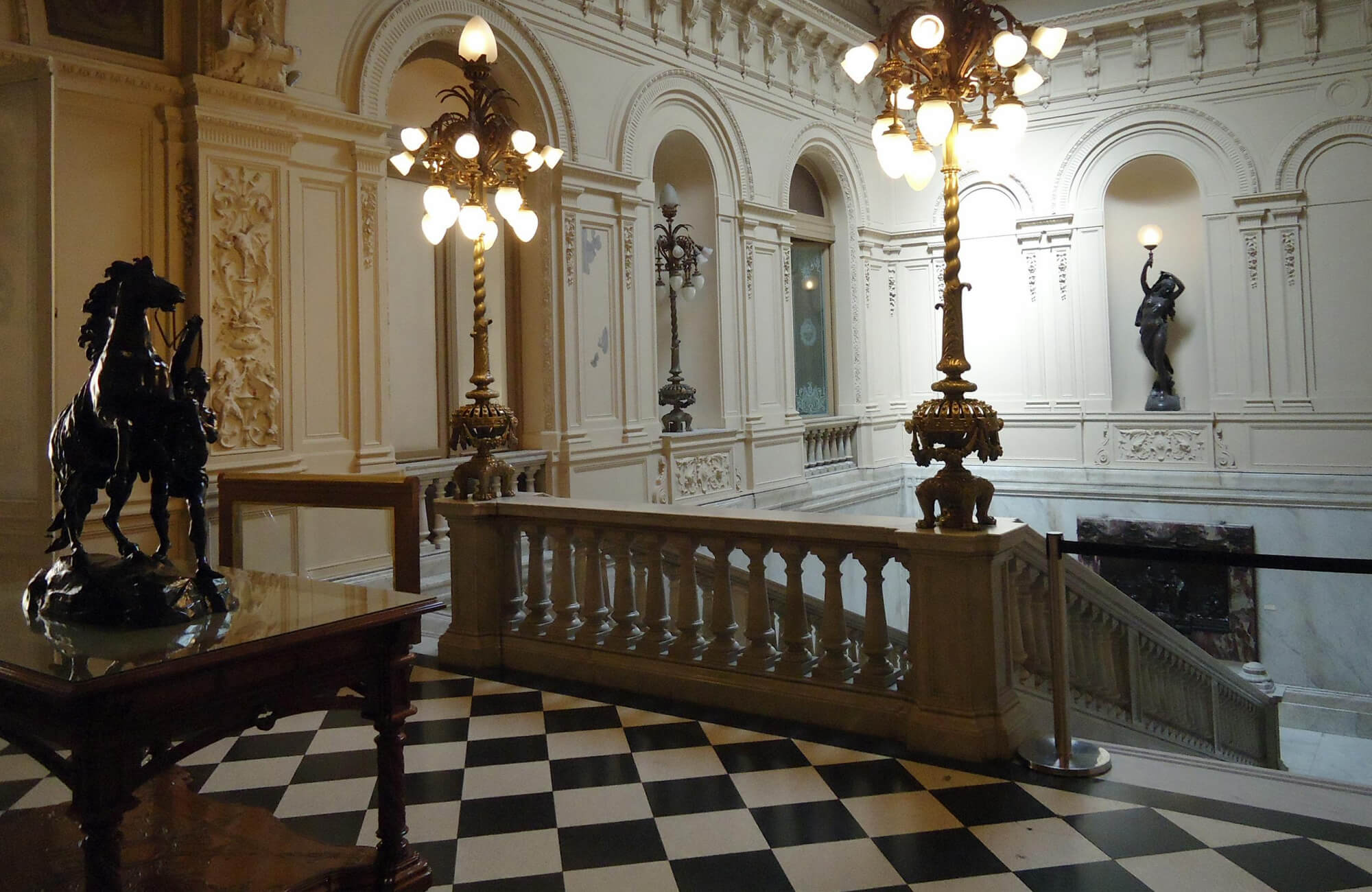 A grand staircase adorned with intricate moldings, gilded lampposts, and a black and white checkerboard floor, exuding classic Beaux-Arts elegance.