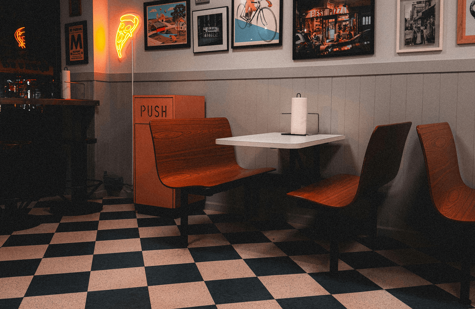 Retro-style diner with a black and white checkerboard floor, wooden booth seating, neon pizza sign, and vintage wall art.