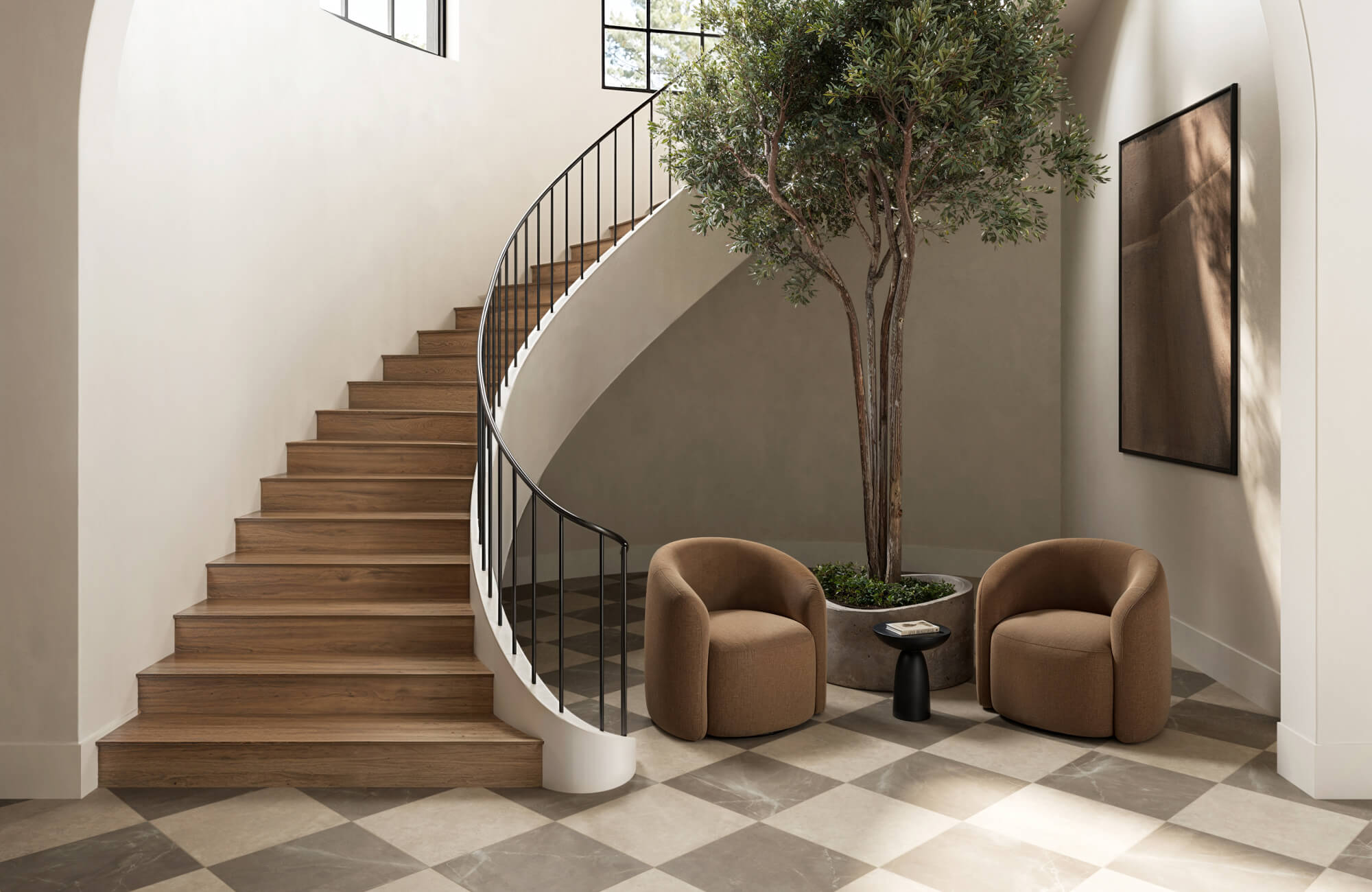 Elegant foyer with a curved staircase, checkerboard floor, earthy-toned seating, and a tall indoor tree adding a natural touch.