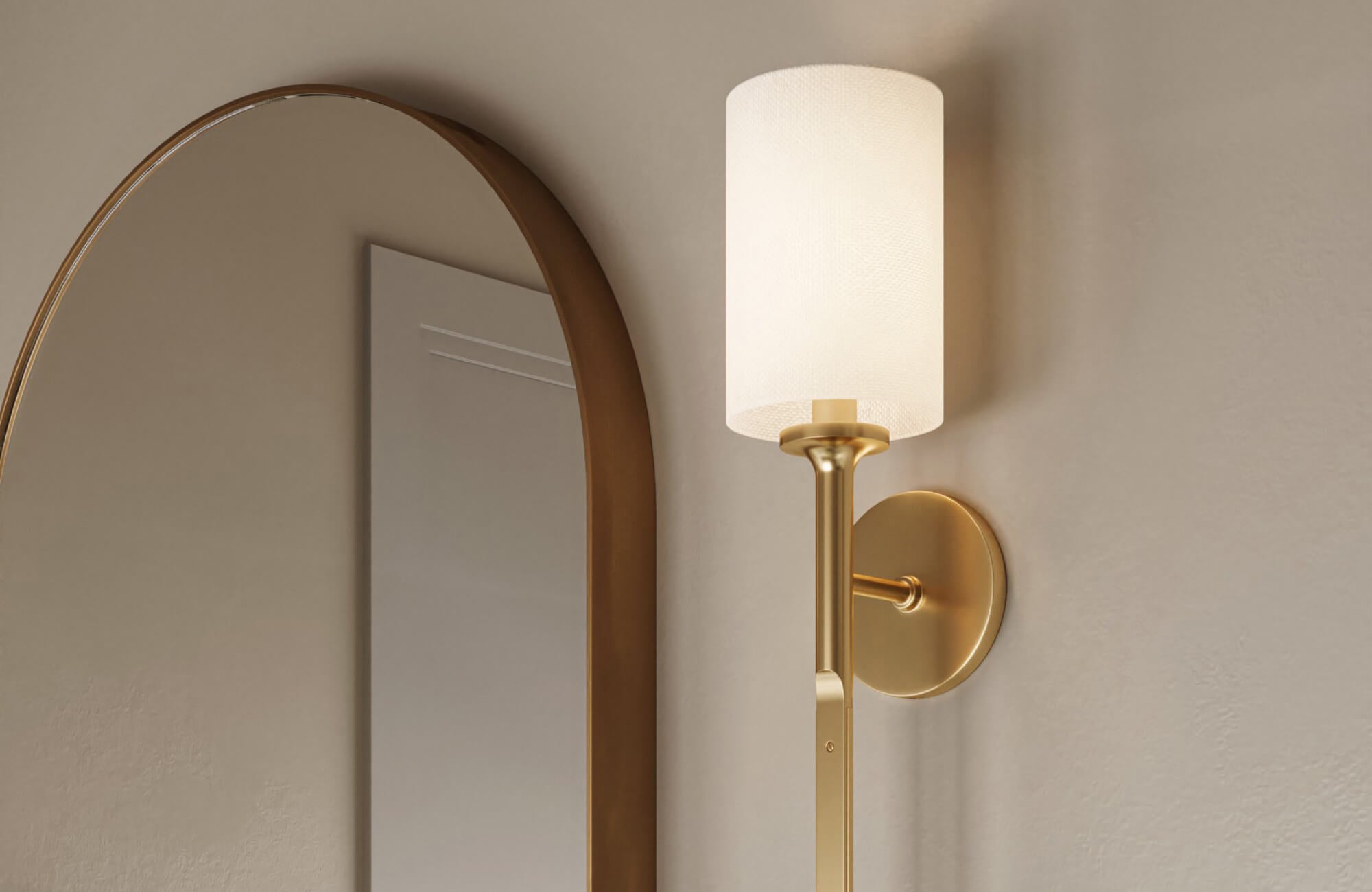 Modern wall sconce with a brushed brass finish and cylindrical white shade mounted beside an arched mirror on a neutral wall.