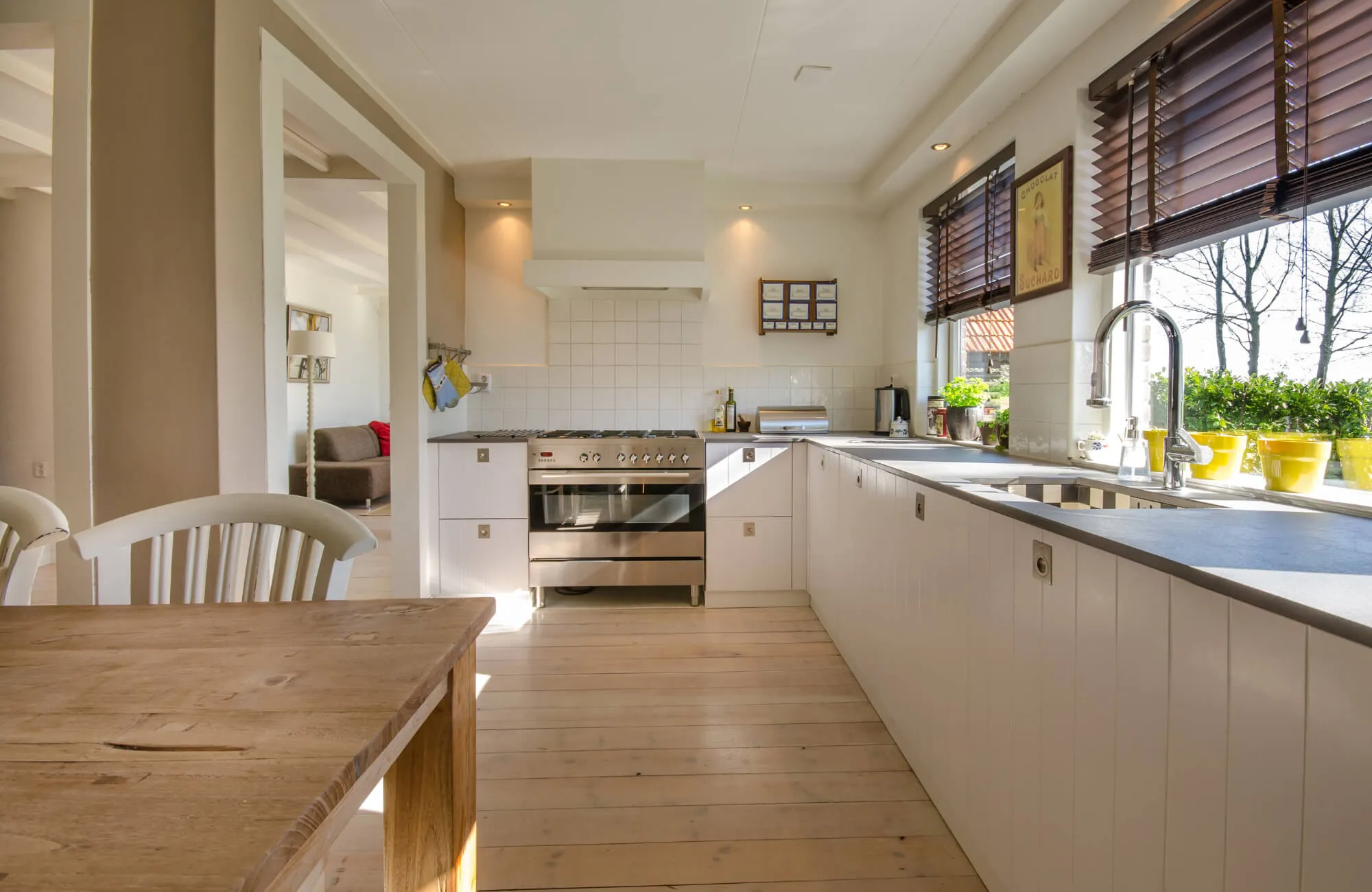 Bright modern kitchen with white cabinets, tiled backsplash, wood flooring, large windows, and stainless steel appliances.