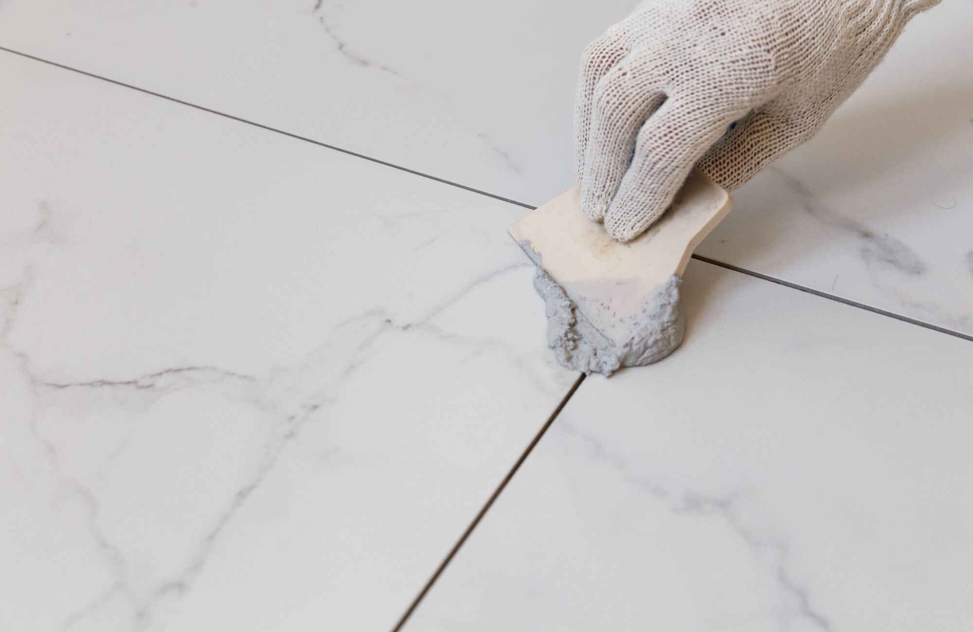 Close-up of a gloved hand applying fresh gray grout between large white marble-effect floor tiles using a plastic spreader tool.