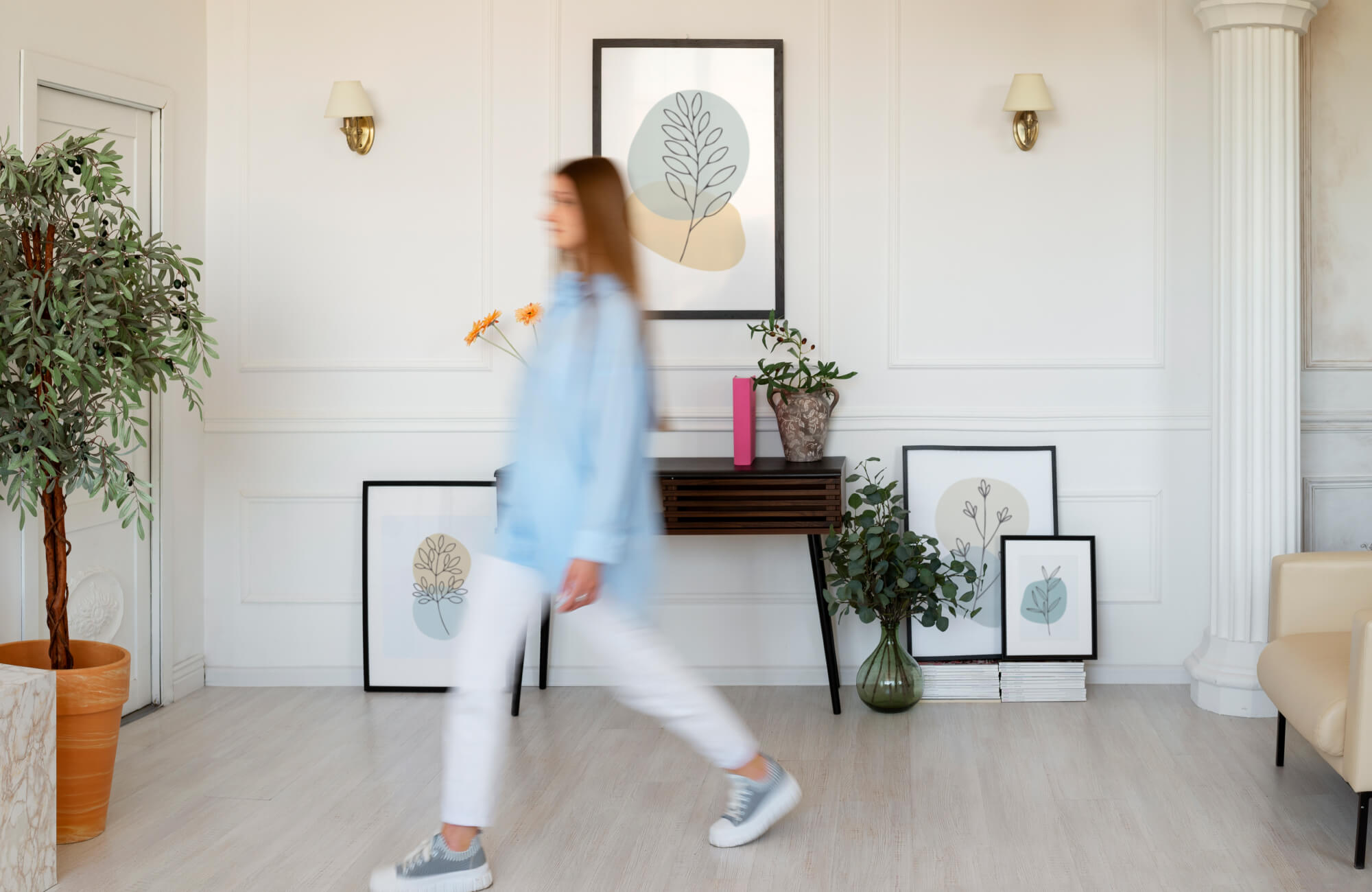 Airy white paneled living space with soft green and beige botanical art prints, a dark brown console table, and a motion-blurred woman in blue passing by.