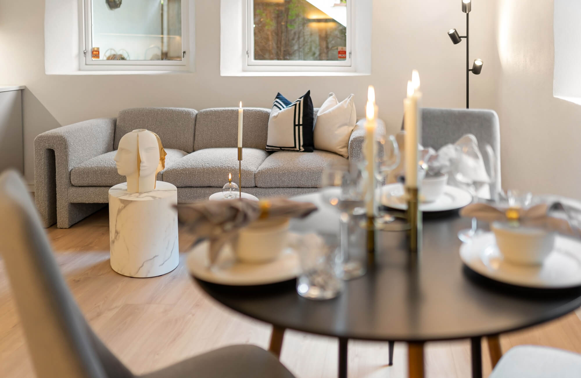 White and gold face bust on a white marble pedestal beside a gray boucle sofa with navy and cream pillows, near a black dining table with gold candles.