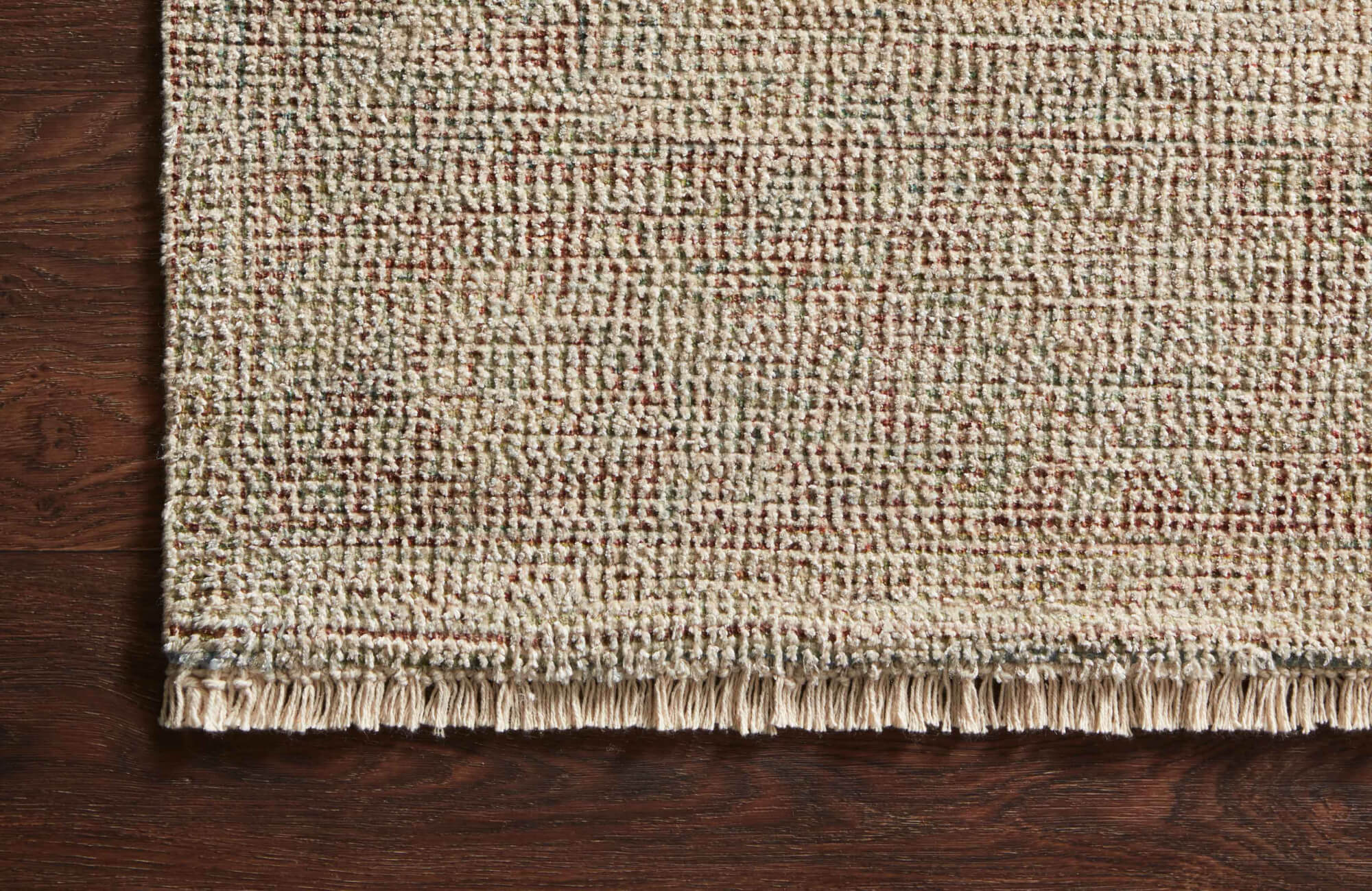 Handwoven non-toxic rug featuring a warm blend of beige, green, and rust threads with knotted fringe, resting on rich brown hardwood.
