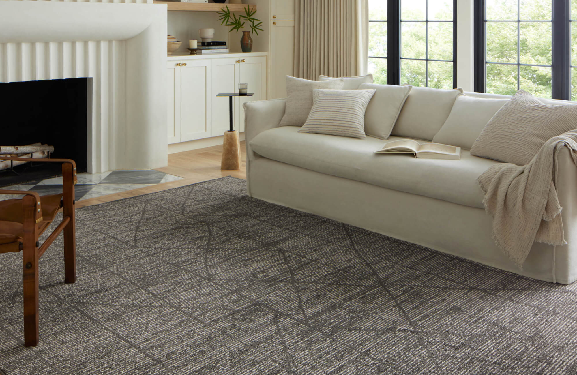Textured gray area rug with organic linear patterns anchors a bright neutral-toned living room with a cream sofa, wood chair, and natural light.