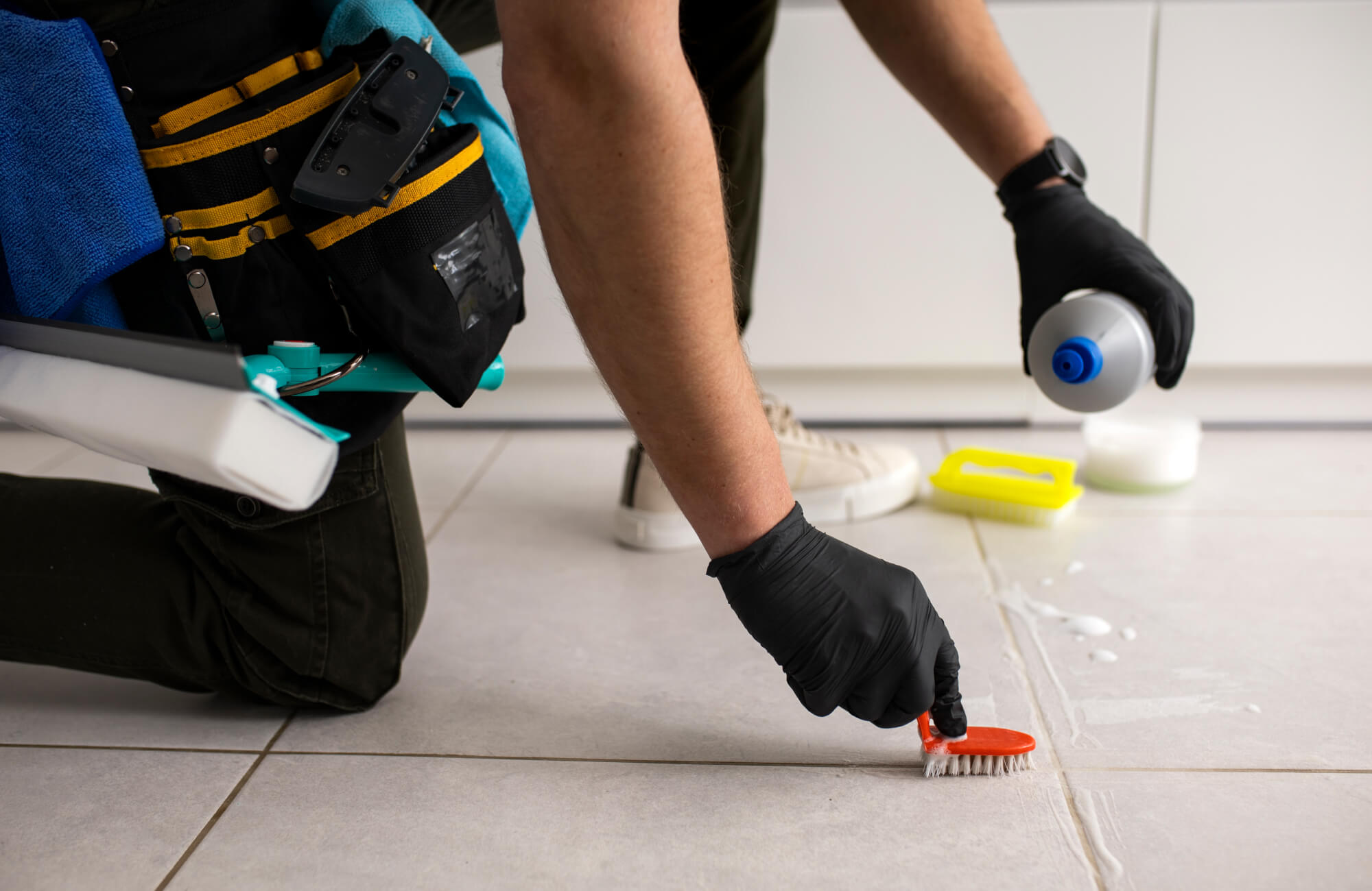 Professional cleaner scrubbing tile grout with a small brush and cleaning solution, equipped with gloves and cleaning tools for precision.