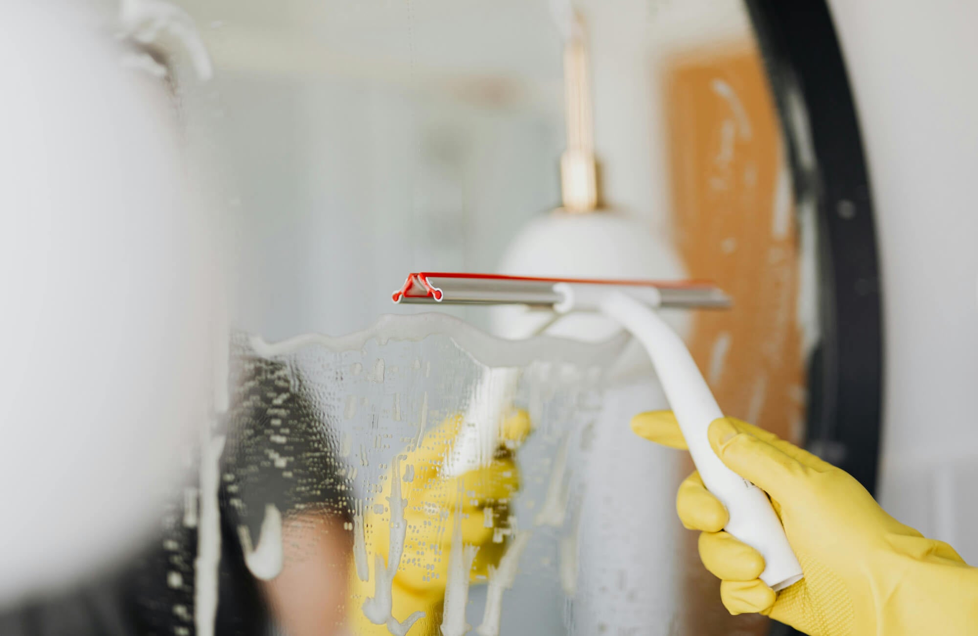 Person wearing yellow gloves cleaning a mirror with a squeegee, removing soap suds for a clear, streak-free finish.