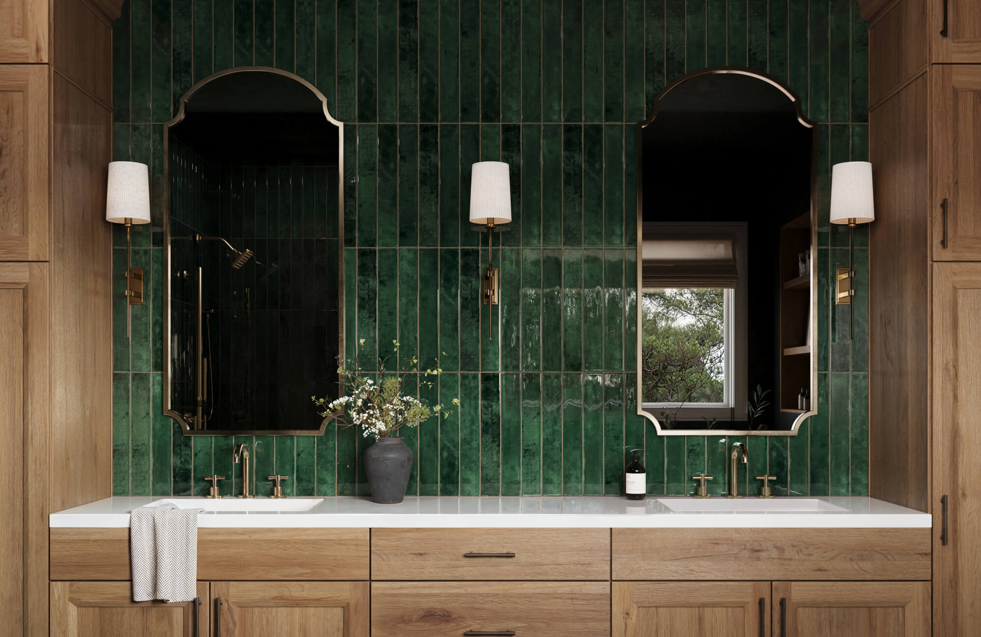 Bathroom vanity with glossy green vertical subway tiles, double sinks, mirrors, wood cabinetry, and brass fixtures.