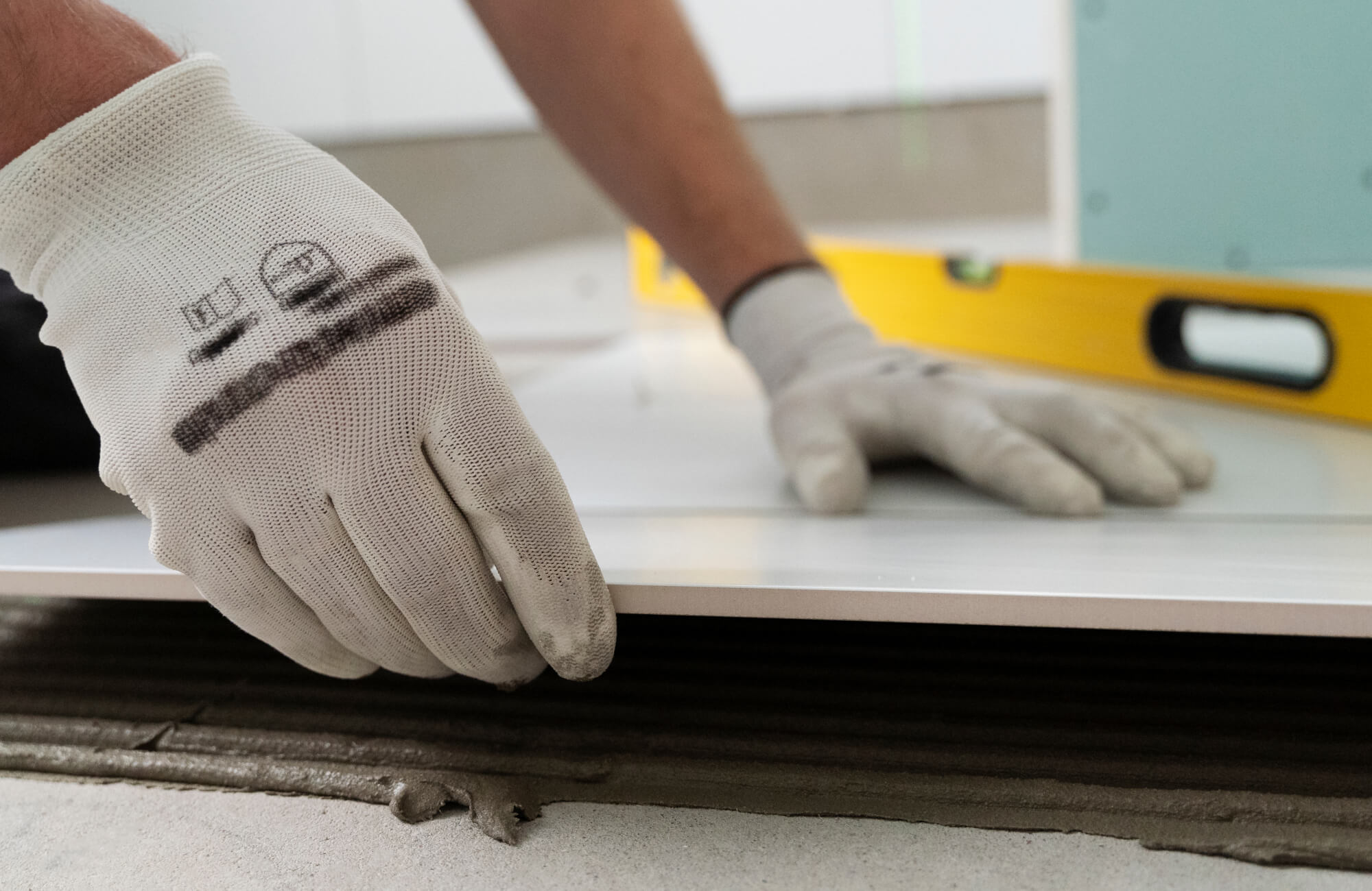 Close-up of tile installation on floor with gloved hands placing large tile over thinset mortar using level for alignment.