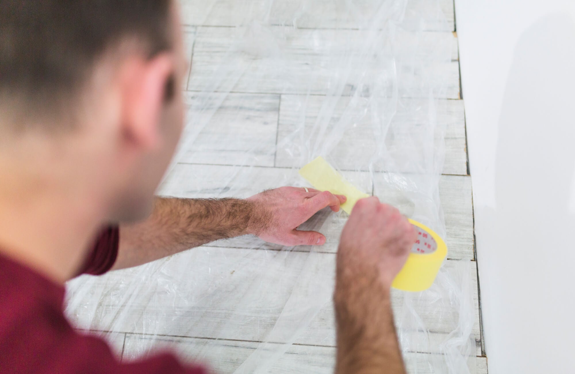 Close-up of floor tiles covered with protective plastic while person tapes surface, showing grout lines and tile layout.