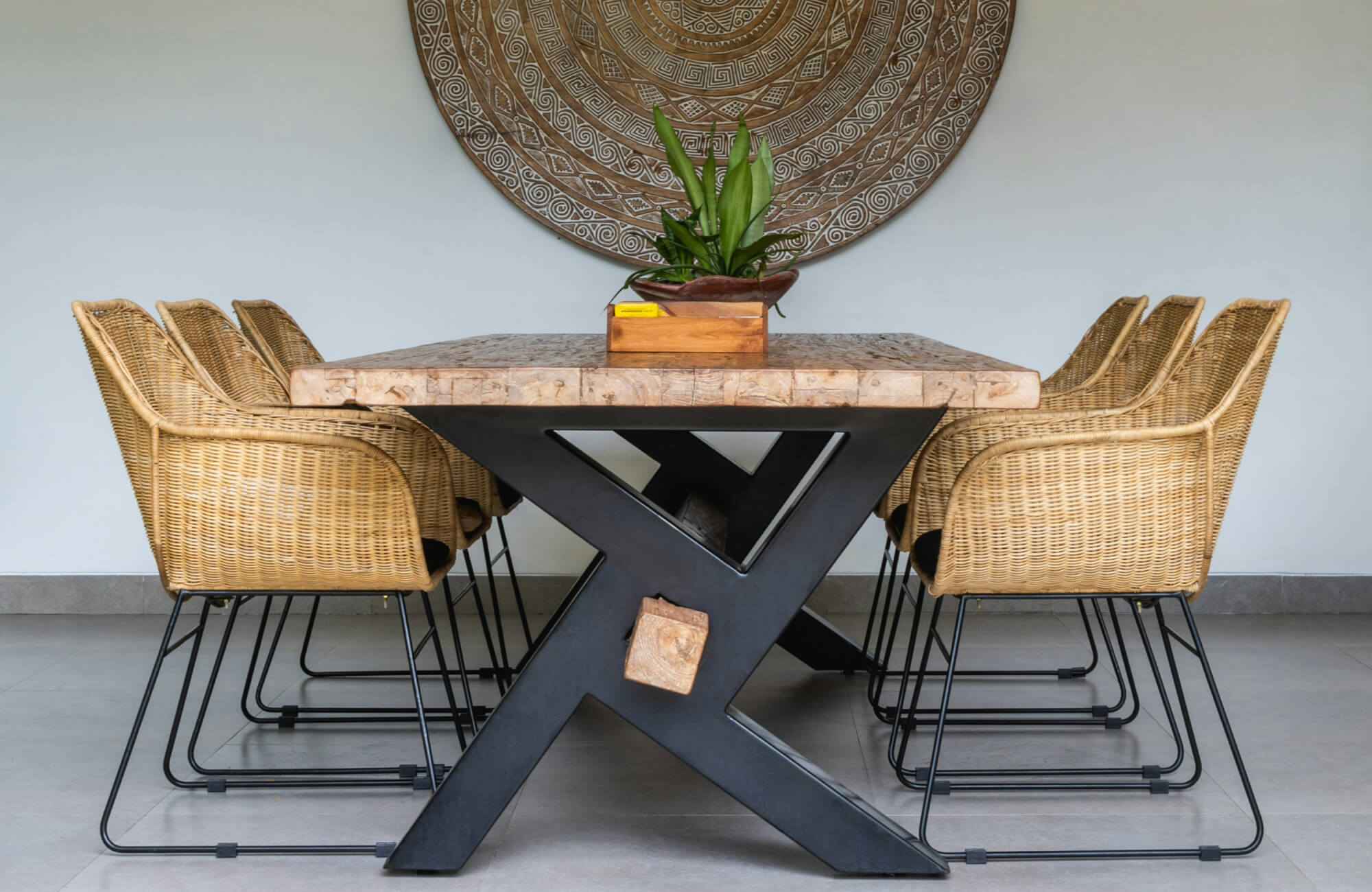 Rustic dining table with thick wood top and black X-base, surrounded by rattan chairs and large carved wall art.