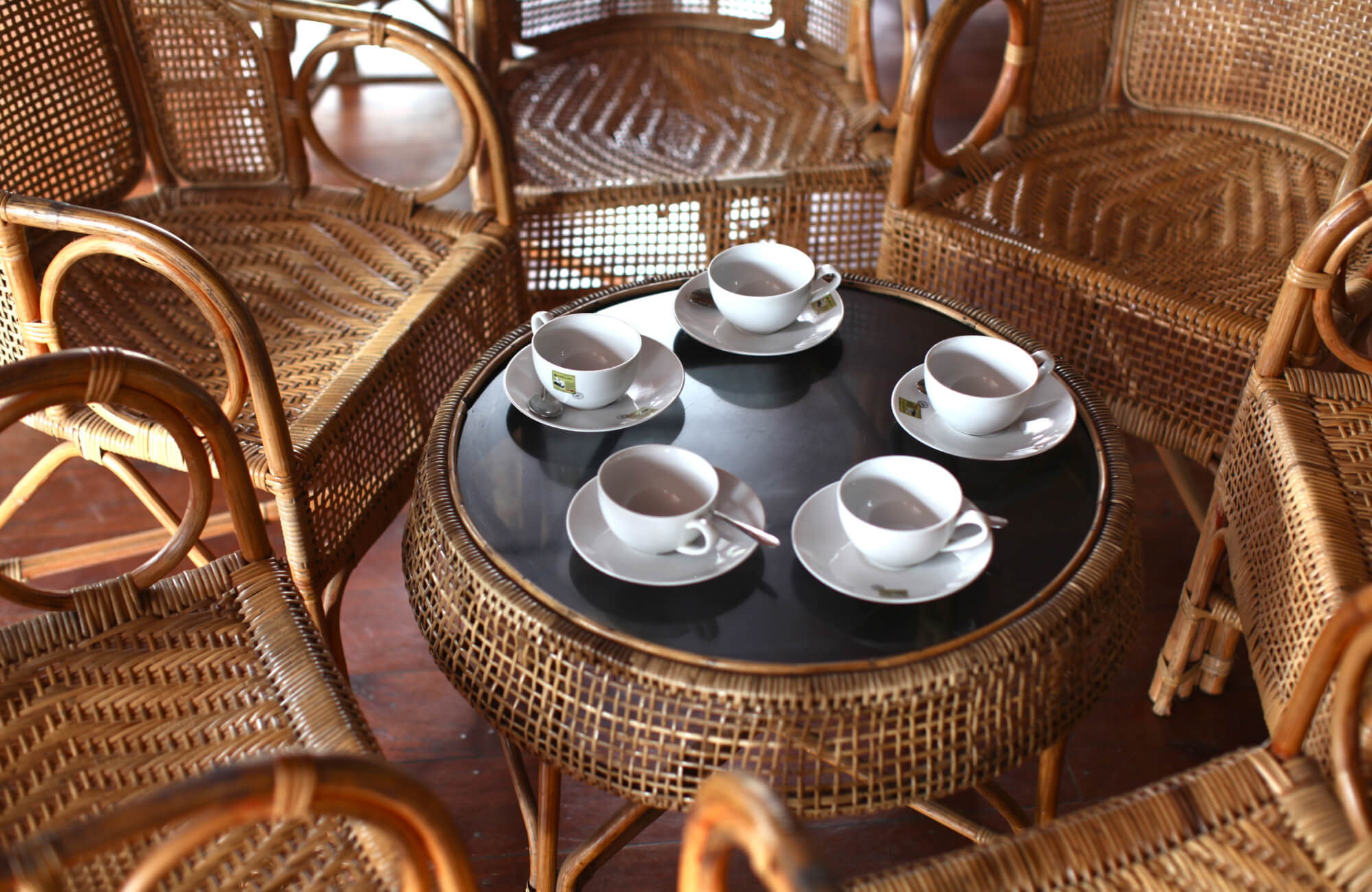 Cozy seating area with a round rattan table and chairs, set with white teacups and saucers on a dark tabletop.