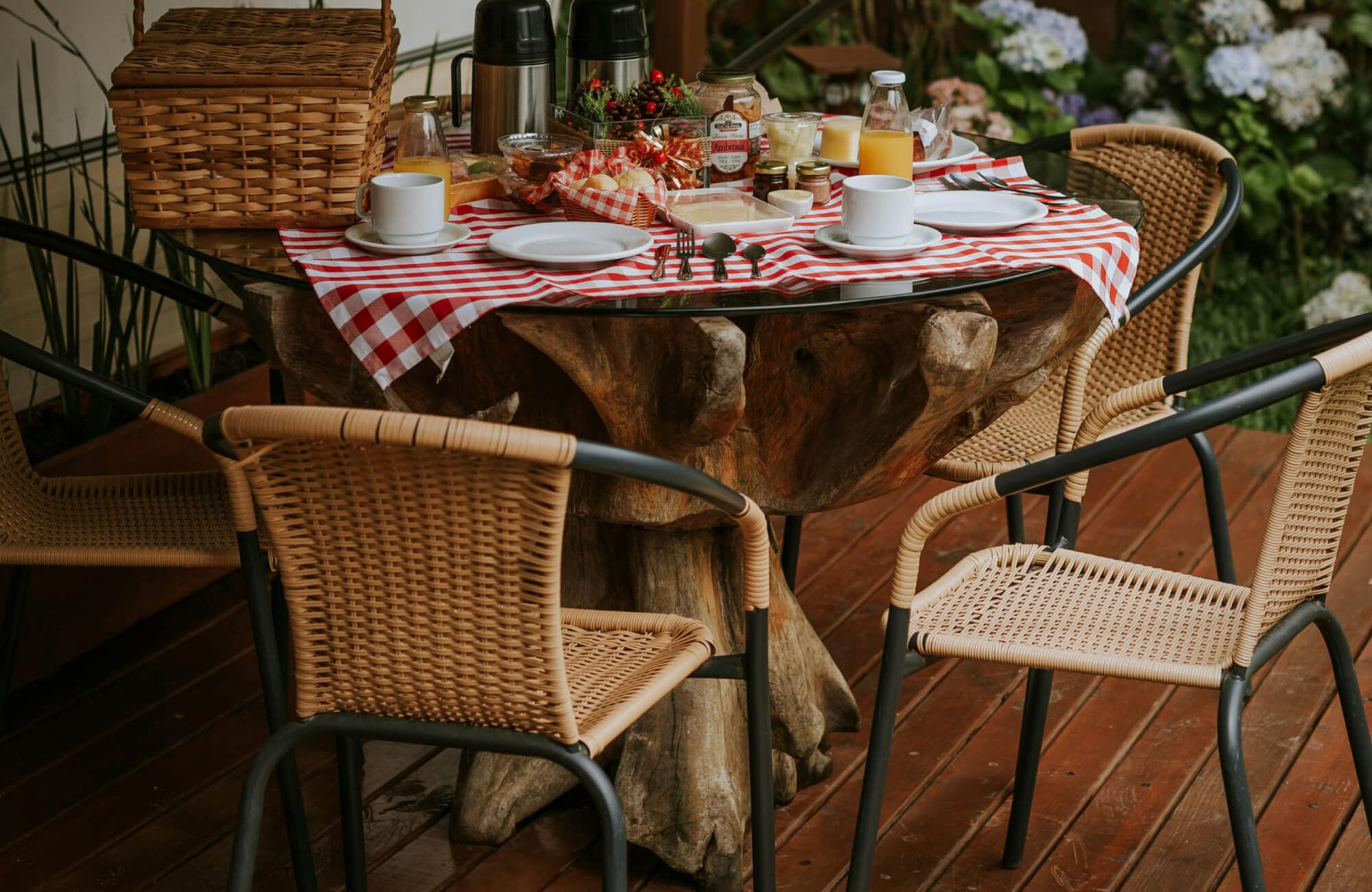 Outdoor patio table with rustic wood base, red gingham tablecloth, breakfast spread, and rattan dining chairs.