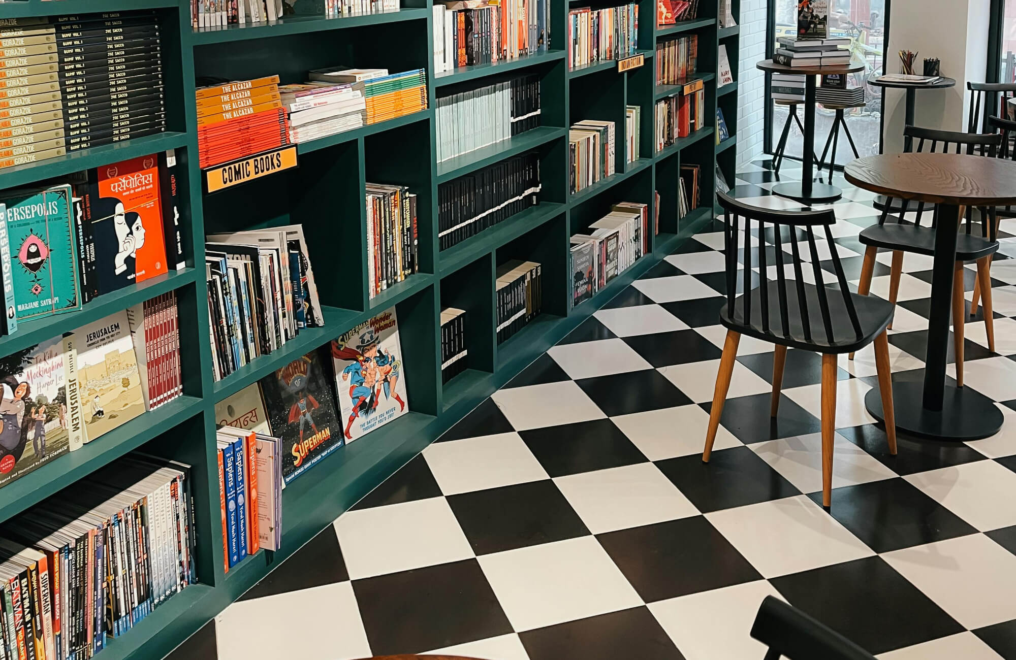 null Cozy bookstore café with black and white checkered tile flooring, teal built-in bookshelves filled with colorful titles, and sleek wooden chairs that create a warm, retro-inspired atmosphere perfect for reading and relaxation.