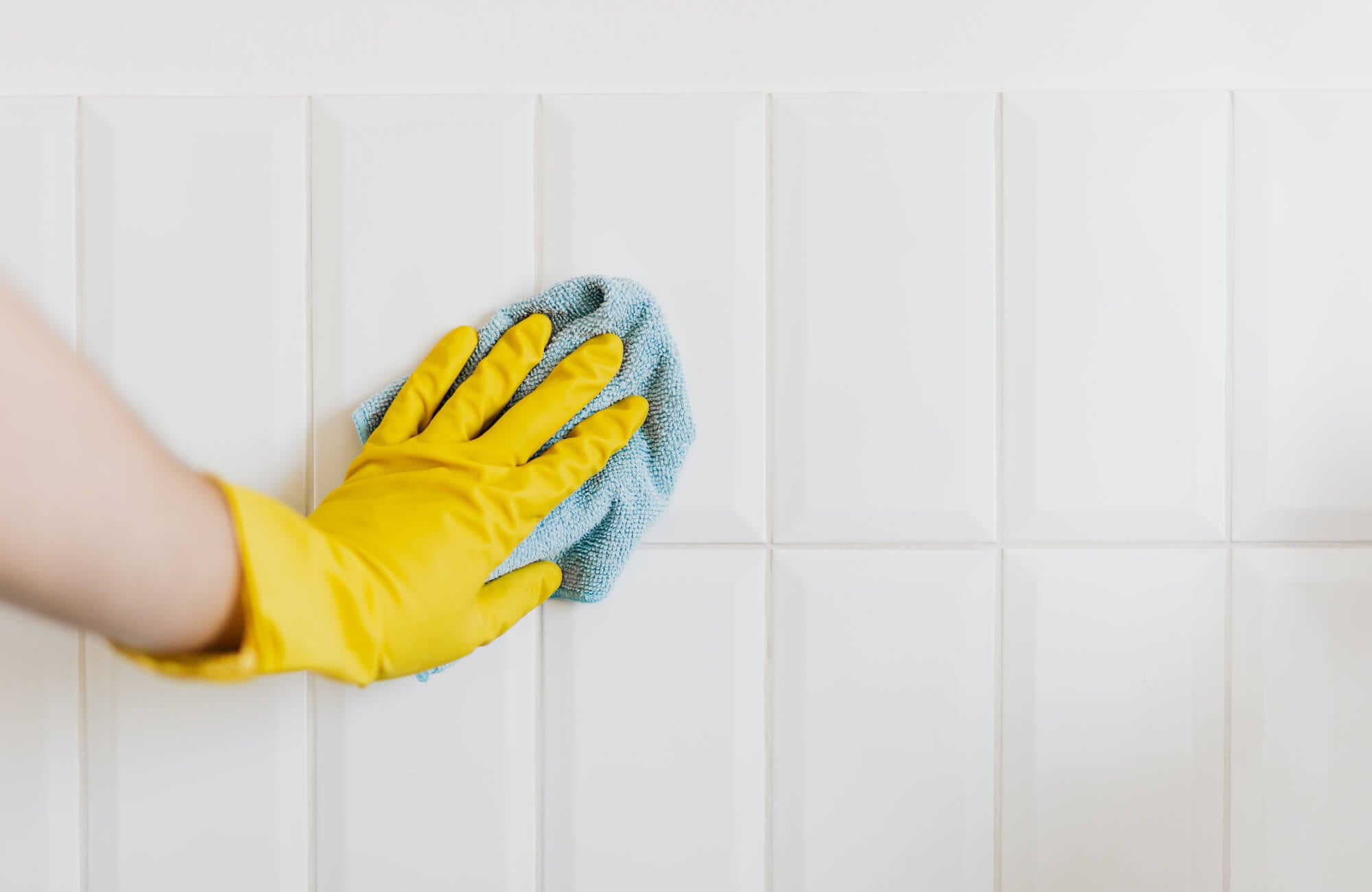 null Hand in yellow glove wiping glossy white ceramic tiles with a blue cloth, illustrating easy kitchen tile cleaning and maintenance.