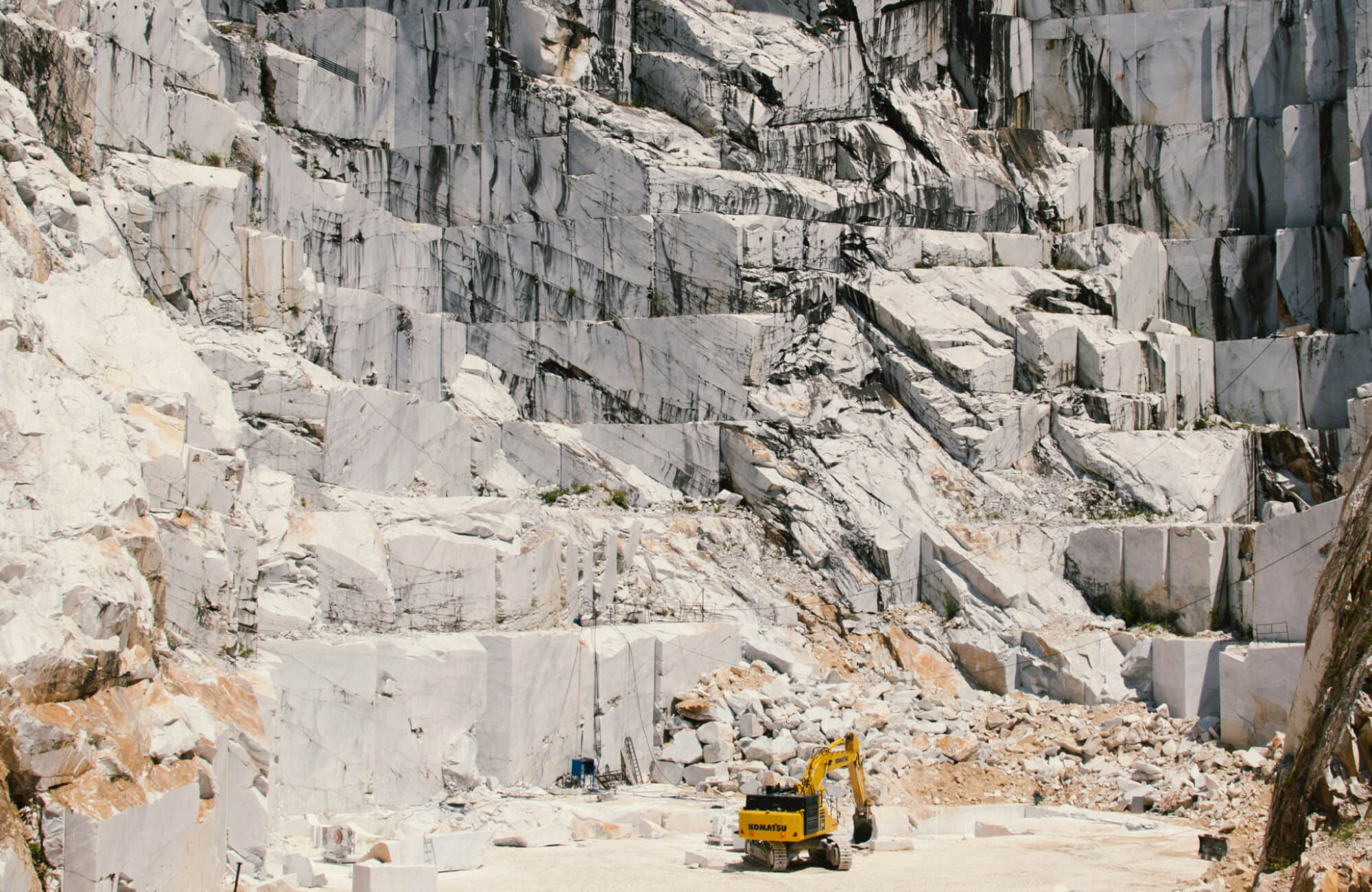 null Active marble quarry with excavator extracting large stone blocks from stepped white rock walls.