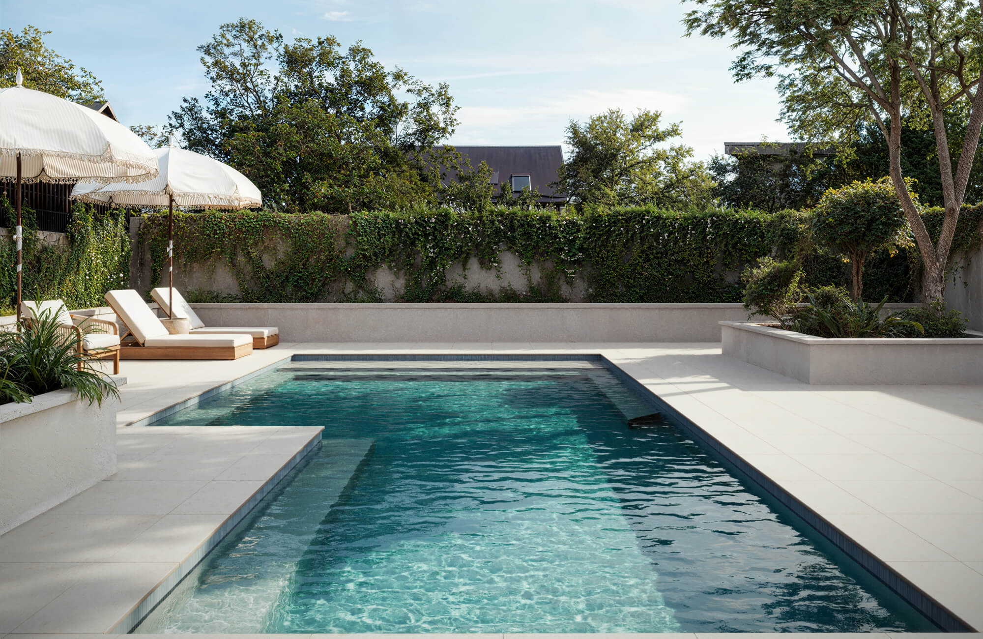 Modern pool lined with slim vertical tiles in muted blue, framed by crisp ivory pavers and lush garden accents.