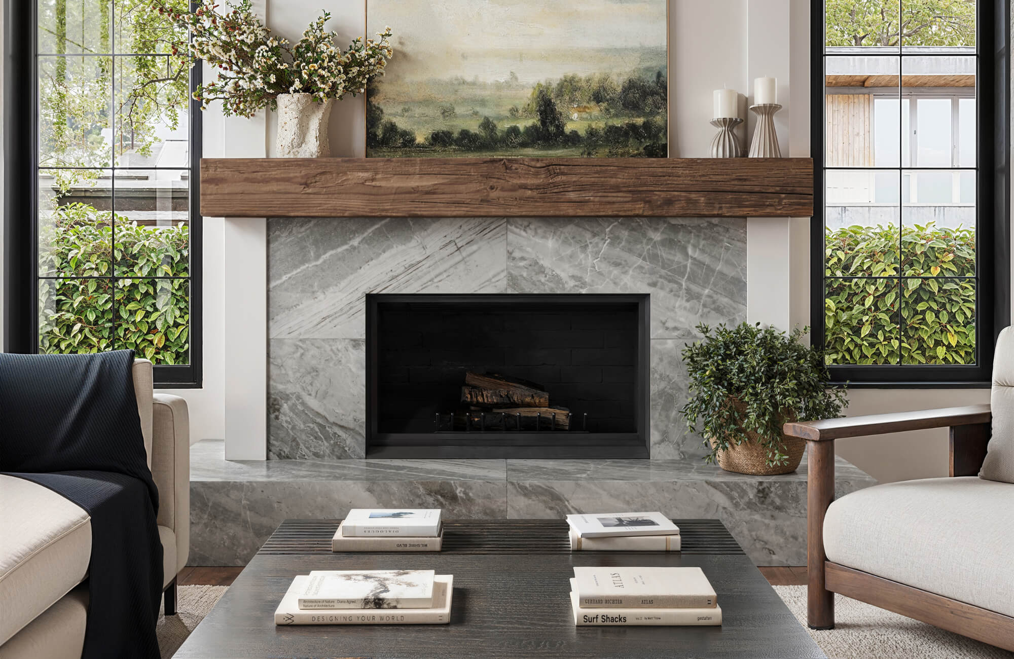 Living room fireplace with polished gray marble look tile surround, thick rustic wood mantel, black firebox, large windows, greenery, and neutral seating.
