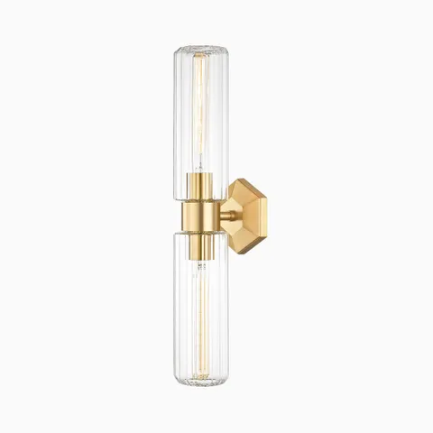 Wall sconce with aged brass finish, hexagonal backplate, and twin vertically fluted glass columns for soft, ambient glow.