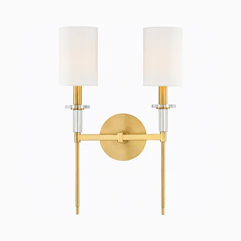Aged brass double wall sconce with two white cylindrical shades, crystal accents, and round backplate for an elegant modern lighting accent.
