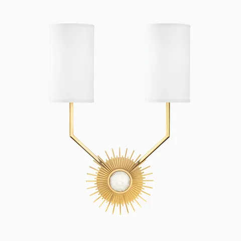 Aged brass wall sconce with dual white cylindrical shades and radiant sunburst backplate, blending bold geometry with modern elegance.