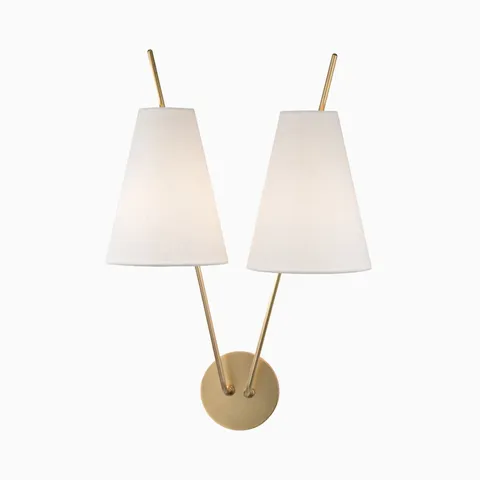 Aged brass wall sconce with round backplate, dual angled arms, and tapered white fabric shades for a modern balanced symmetrical design.