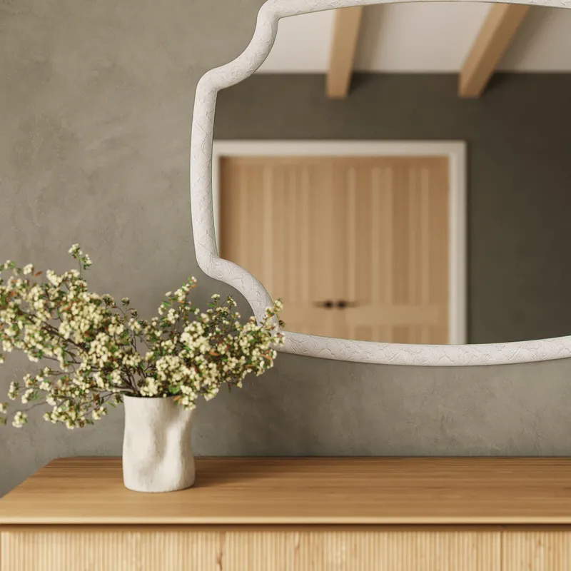 White textured Seraphin mirror with sculpted resin frame displayed above a wood dresser, reflecting warm ceiling beams in a serene interior.