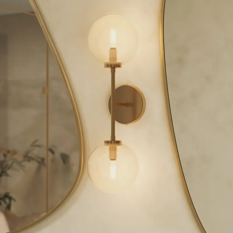Modern bathroom wall sconce with dual clear glass globe shades and brushed brass arm beside an asymmetrical mirror.
