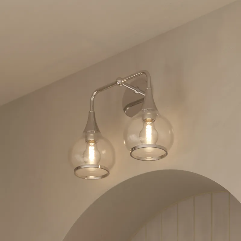 Chrome double wall sconce with curved arm and clear glass globe shades, illuminated above an arched mirror.