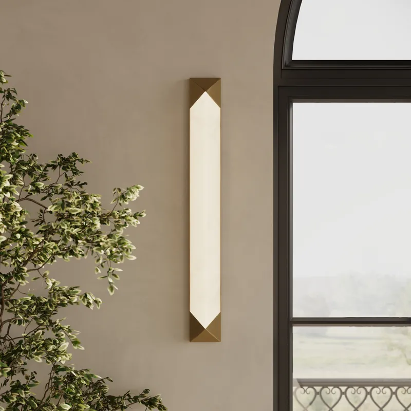 Modern vertical LED wall sconce with alabaster-style diffuser and geometric brushed brass caps mounted beside an arched window.