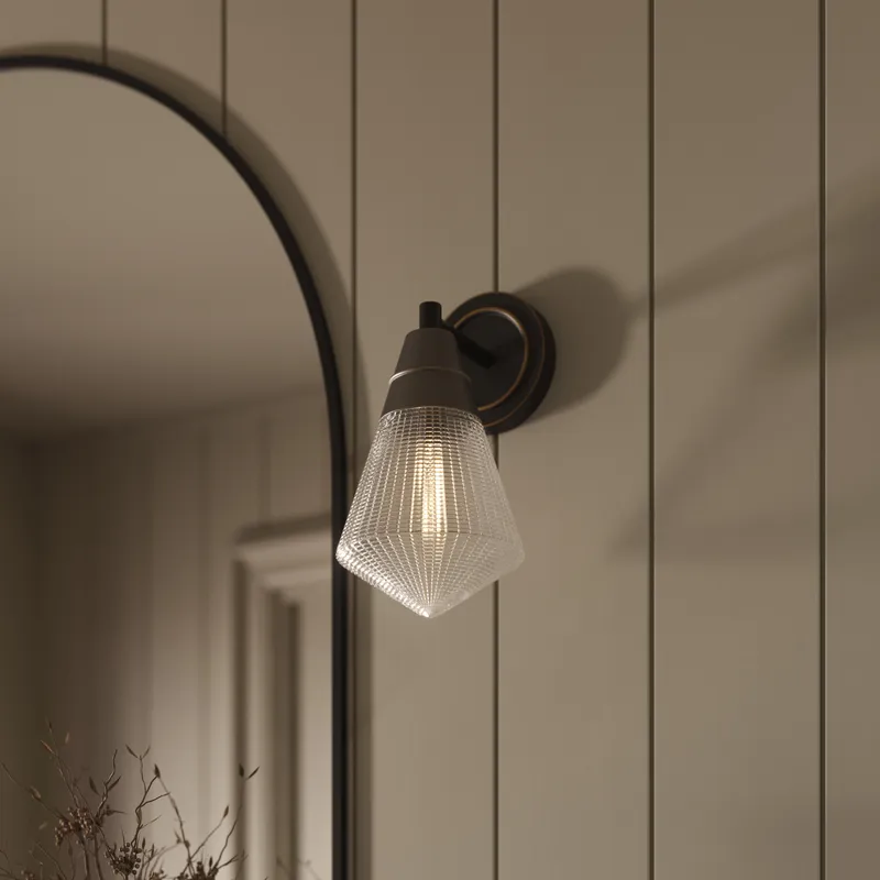 Matte black wall vanity light with a faceted glass shade softly lighting the space beside a curved mirror on a light paneled wall.