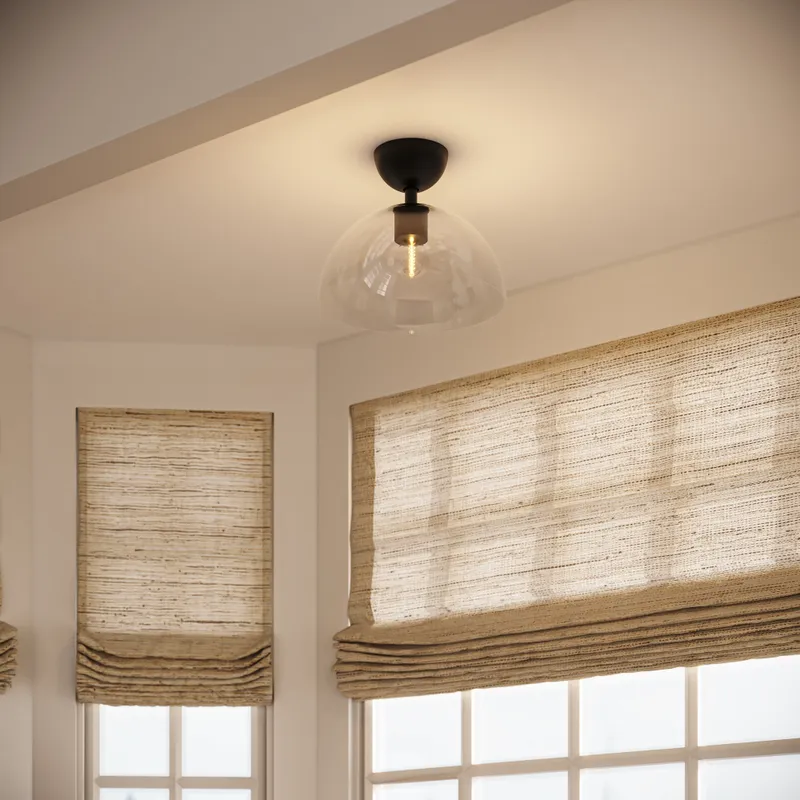 Matte black semi flush mount ceiling light with a clear glass dome shade installed above corner windows with woven Roman shades, casting a warm glow across the soft neutral walls.